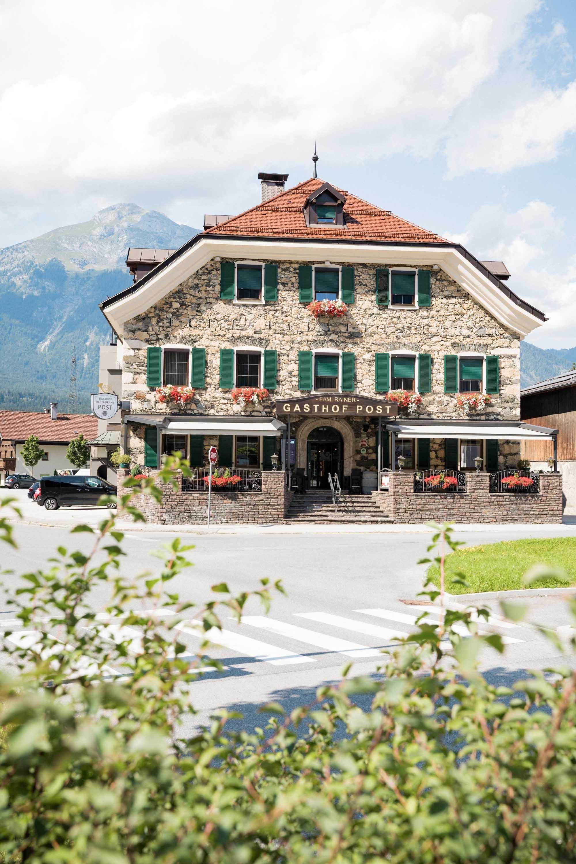 Ein schönes, traditionelles Gasthaus mit grünen Fensterläden und Blumen. Im Hintergrund sind Berge und ein blauer Himmel zu sehen.
