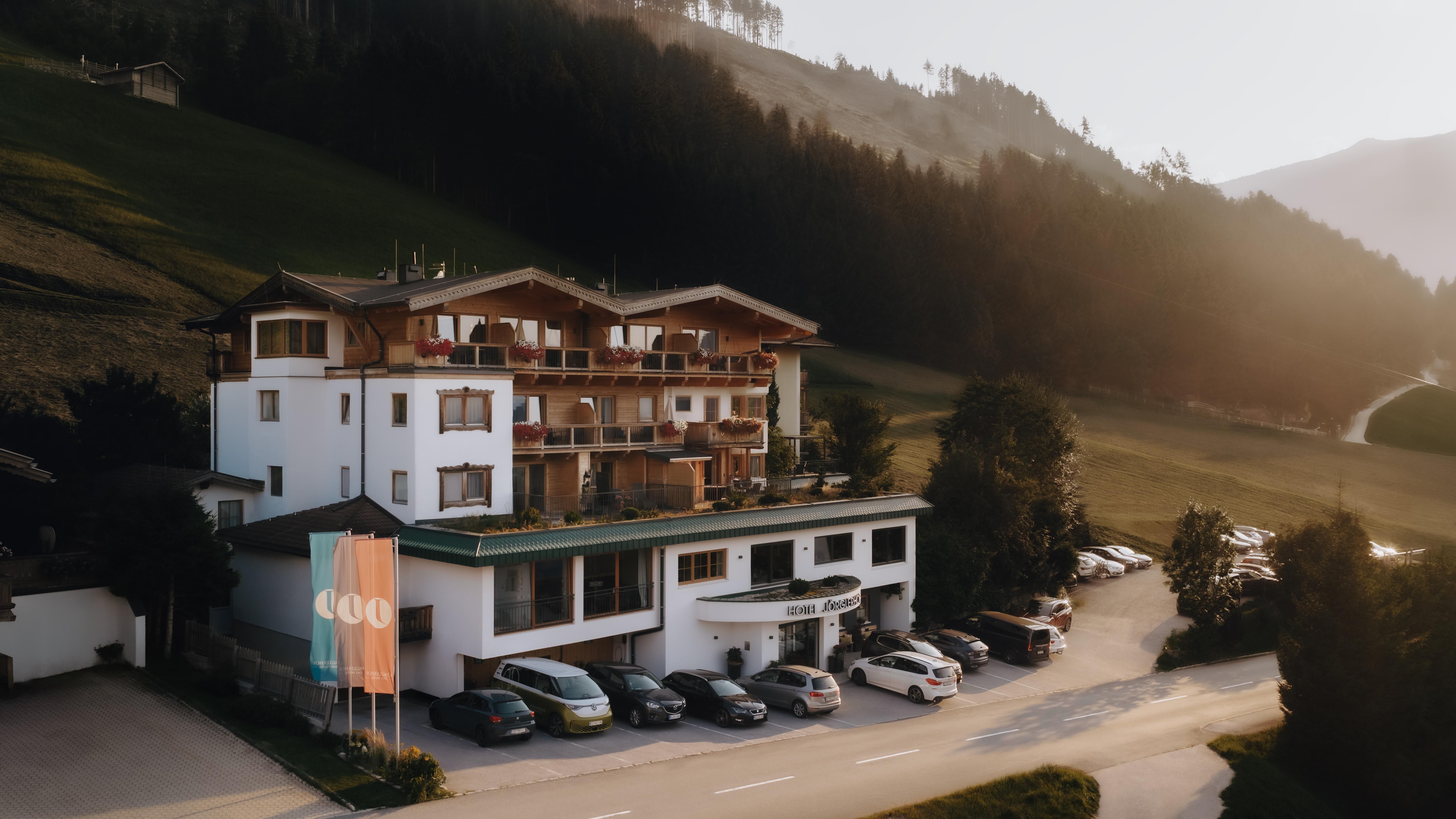 Ein gemütliches Hotel in den Bergen mit mehreren Balkonen und großen Fenstern. Im Vordergrund stehen geparkte Autos und eine ruhige Straßenlandschaft.