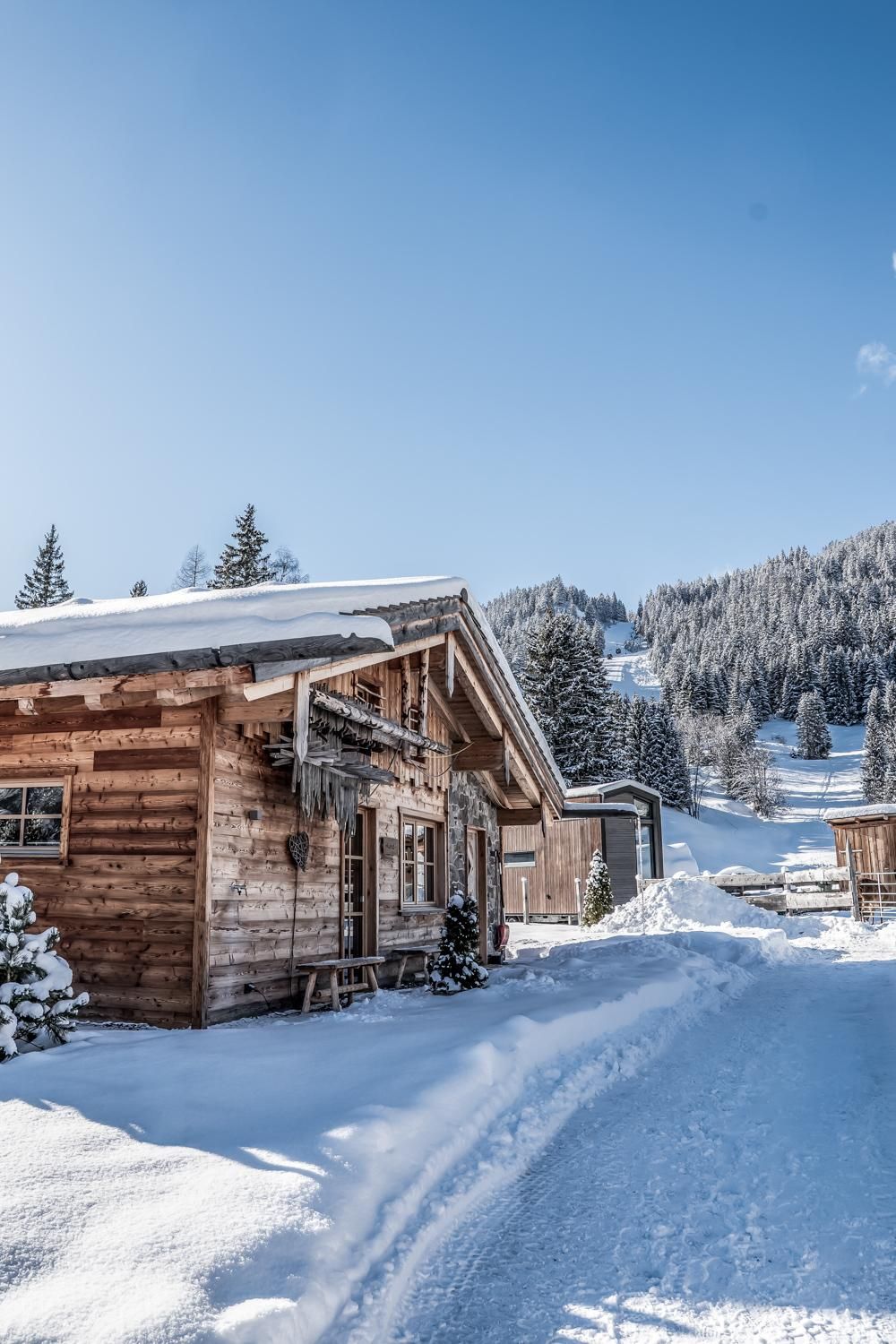 Ein winterliches Chalet in einer verschneiten Landschaft. Im Hintergrund erheben sich majestätische Berge unter einem klaren blauen Himmel.