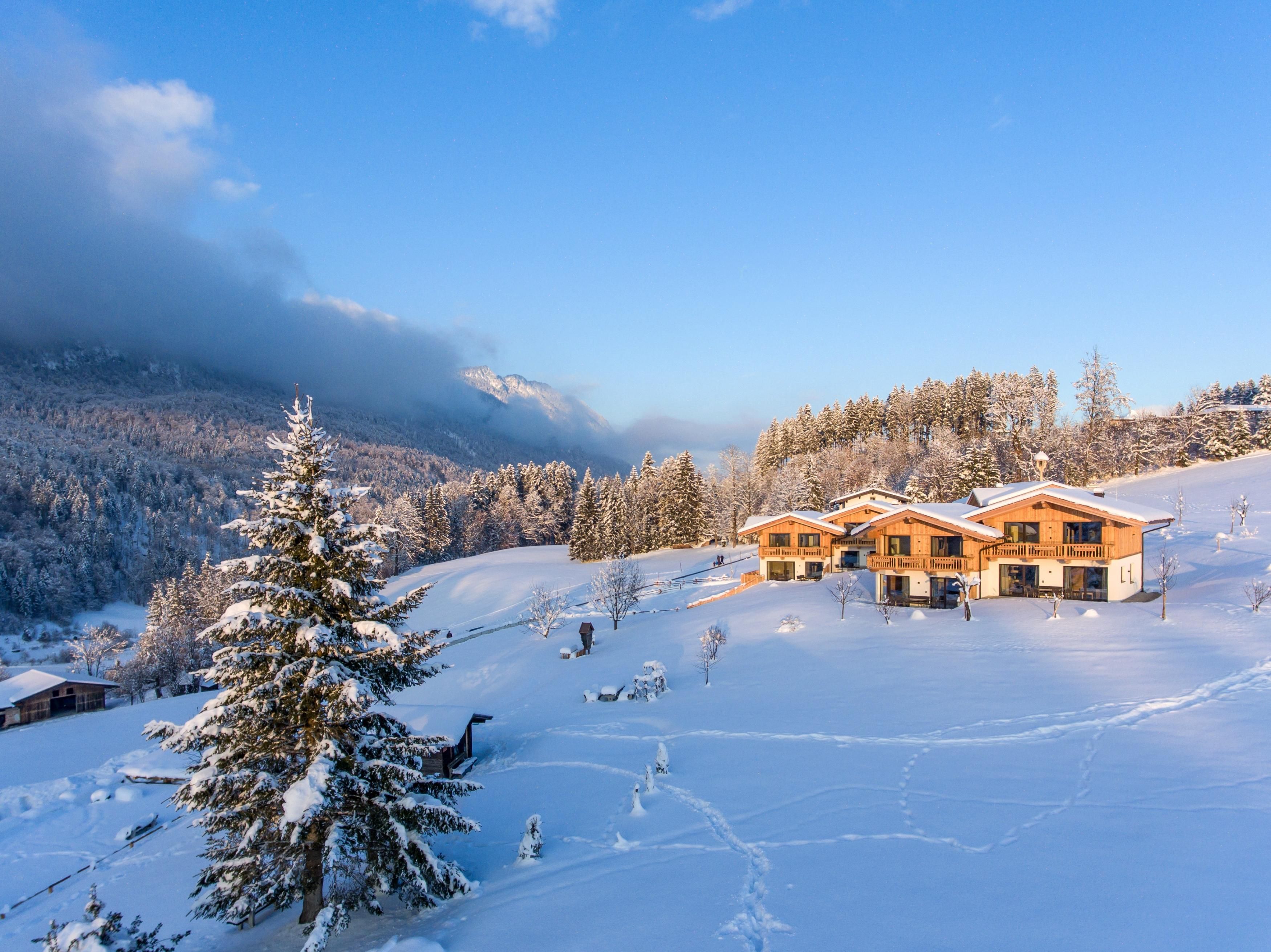 Eine winterliche Landschaft mit schneebedeckten Hügeln und drei gemütlichen Holzhäusern. Im Hintergrund sind Wälder und der blaue Himmel zu sehen.