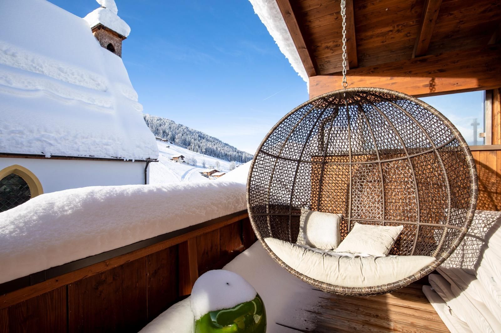 Ein gemütlicher Hängestuhl auf einer Balkonterrasse, umgeben von Schnee. Im Hintergrund sind schneebedeckte Berge und ein klarer blauer Himmel zu sehen.