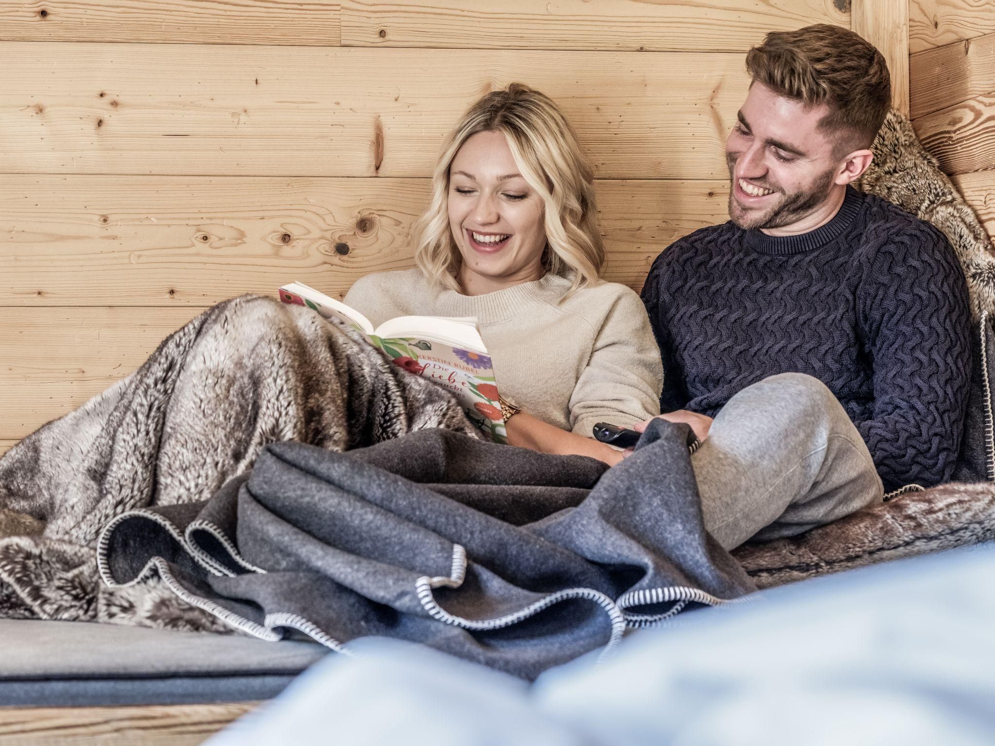 Ein Paar sitzt gemütlich in einem Chalet und liest zusammen ein Buch. Sie sind mit einer Decke zugedeckt und lächeln entspannt.