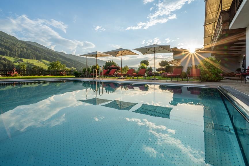 Ein schön gestalteter Pool in den Alpen, umgeben von grünen Hügeln. Sonnenstrahlen scheinen durch die Wolken und reflektieren sich im Wasser.