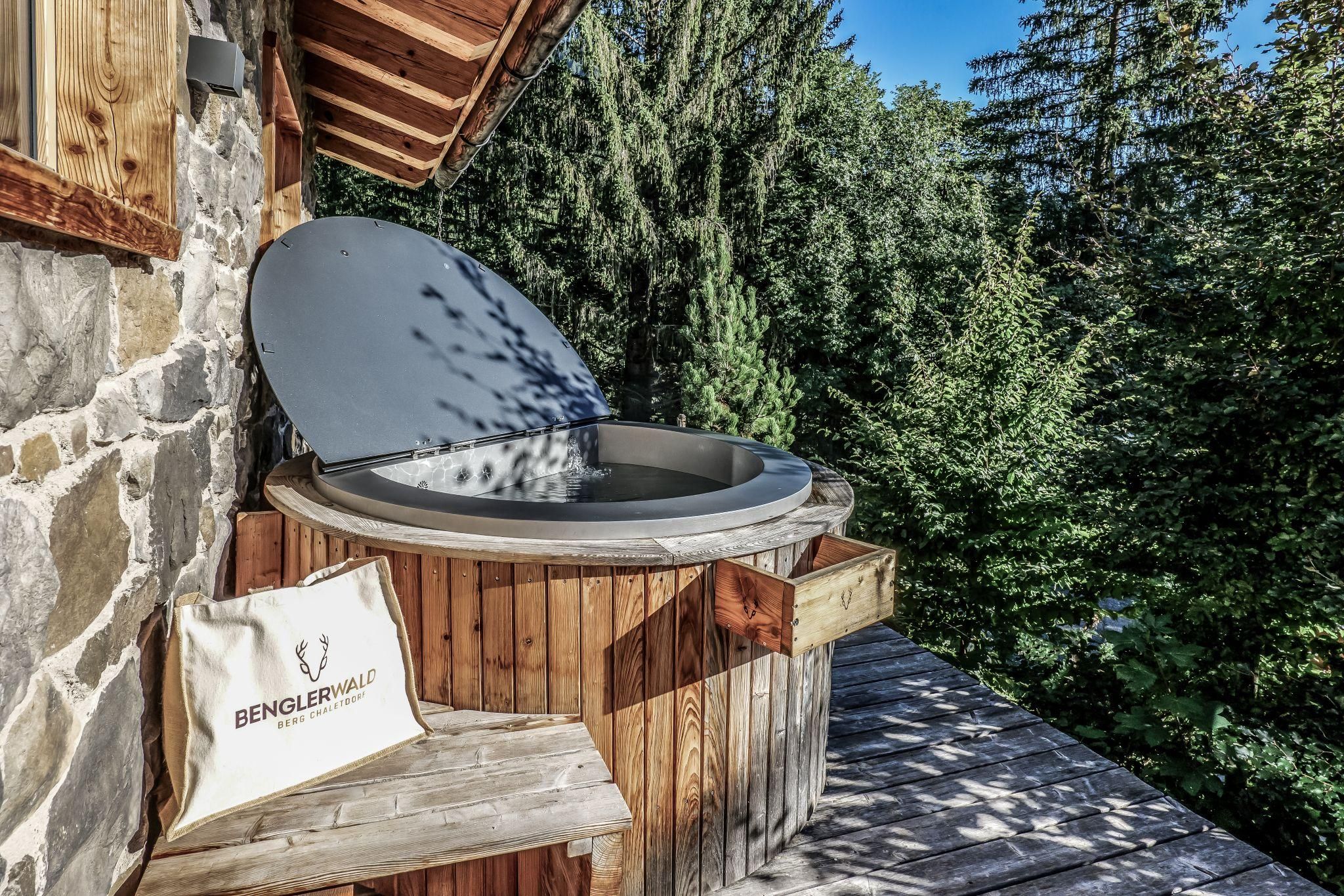 Ein Entspannungsbereich mit einem modernen Hot Pot auf einer Holzterrasse. Umgeben von grünen Bäumen und blauem Himmel bietet es eine ruhige Atmosphäre.