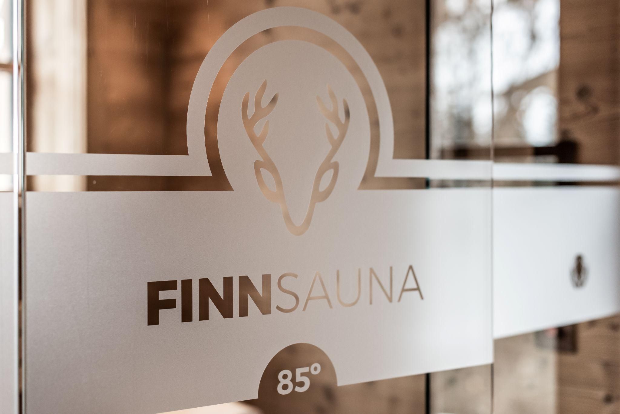 Ein klares Glas mit dem Schriftzug "FINN SAUNA" und einem Hirschlogo. Daneben befindet sich eine Holzstruktur, die eine entspannende Atmosphäre schafft.