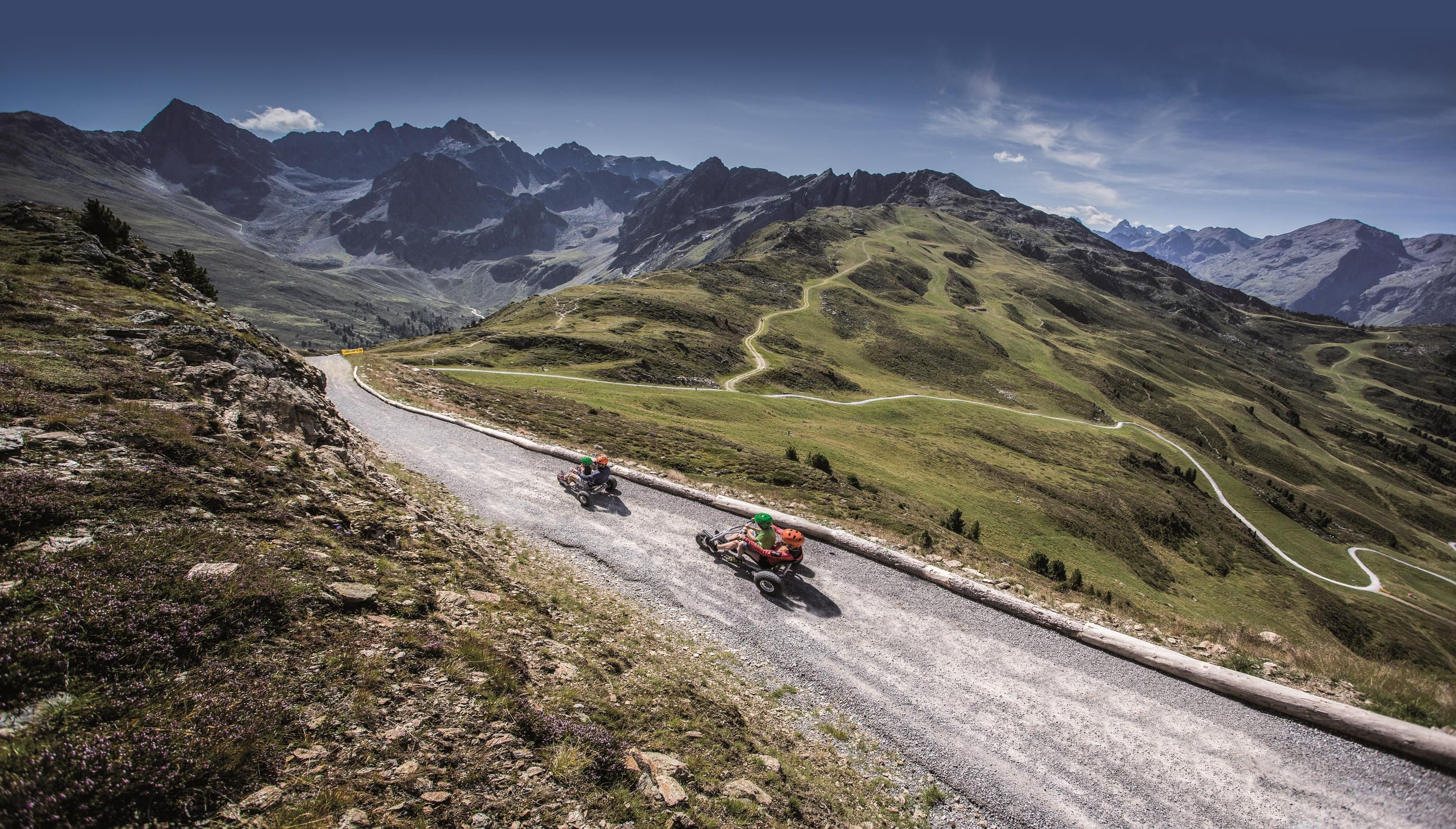 Eine malerische Landschaft mit grünen Hügeln und schroffen Bergen im Hintergrund. Zwei Fahrer auf Motorrädern fahren eine kurvenreiche Straße entlang.