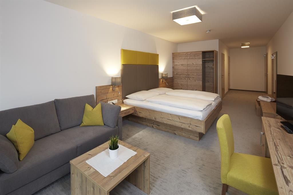 Ein modernes Zimmer mit einem Doppelbett, einer grauen Couch und einem Holztisch. Die Einrichtung ist hell und freundlich, mit bunten Akzenten.