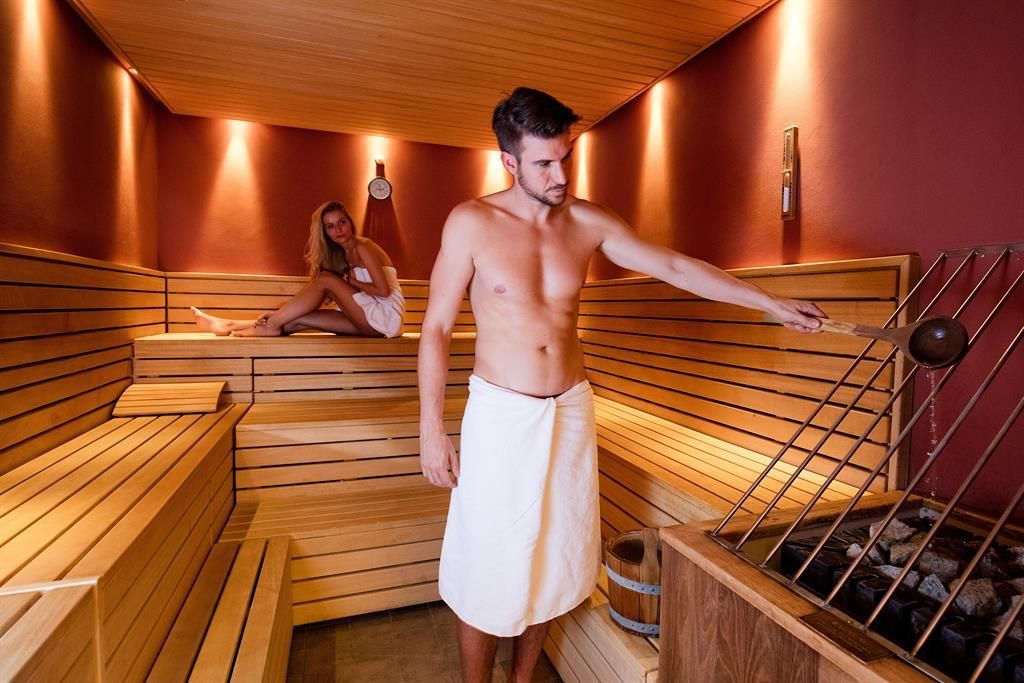 Eine moderne Sauna mit Holzverkleidung. Ein Mann gießt Wasser auf die heißen Steine, während eine Frau entspannt auf einer Bank sitzt.