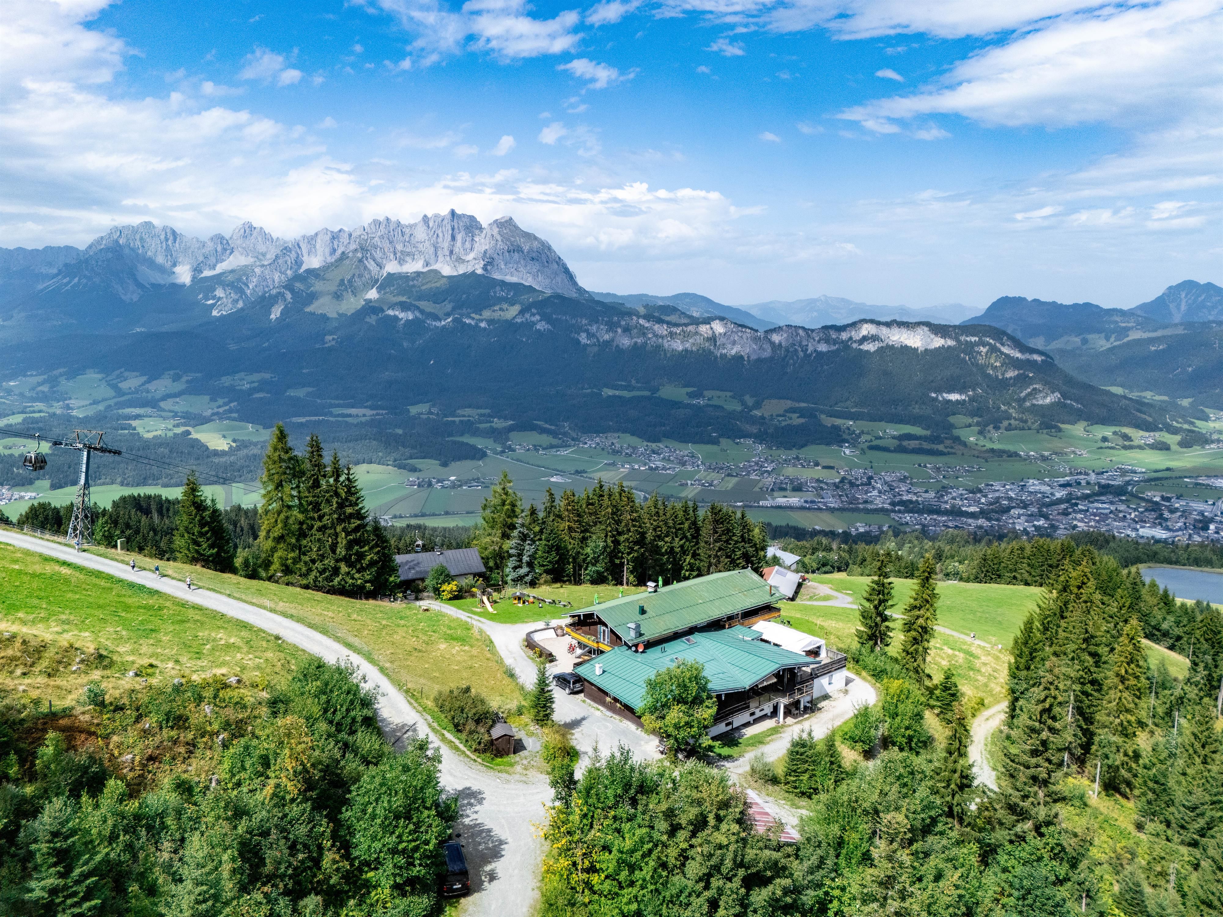 Eine malerische Berglandschaft mit hohen Gipfeln und einer grünen Wiese. Im Vordergrund steht eine gemütliche Berghütte umgeben von Tannen.