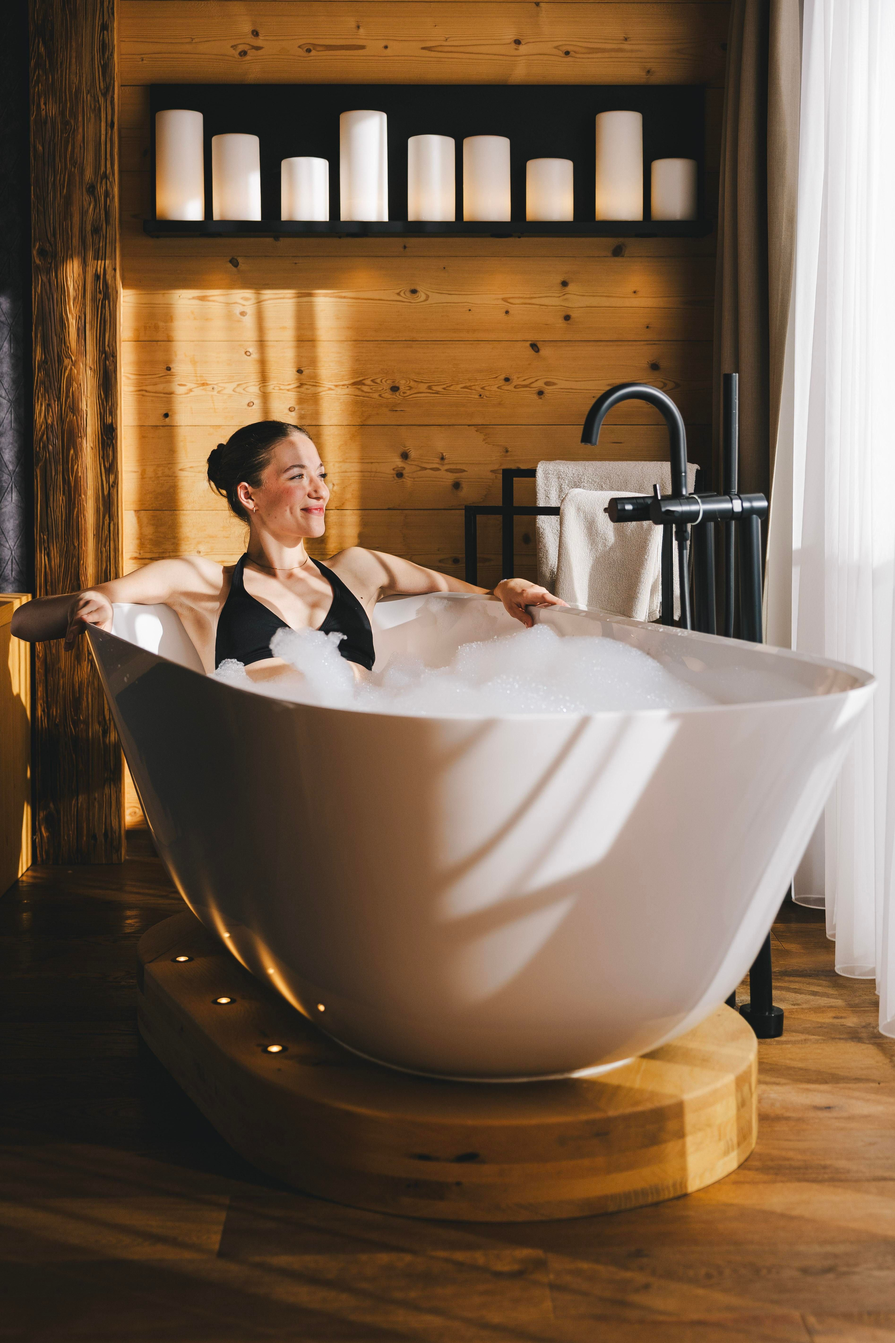 Eine Frau entspannt in einer modernen Badewanne mit Schaum. Der Raum hat warmes Holz und sanfte Beleuchtung.