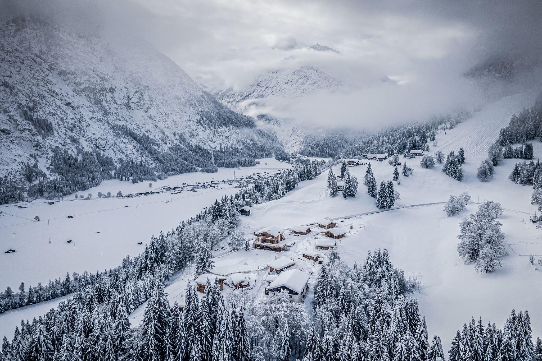 Eine verschneite Landschaft mit Chalets und dichten Bäumen. Die umliegenden Berge sind von Wolken umhüllt.