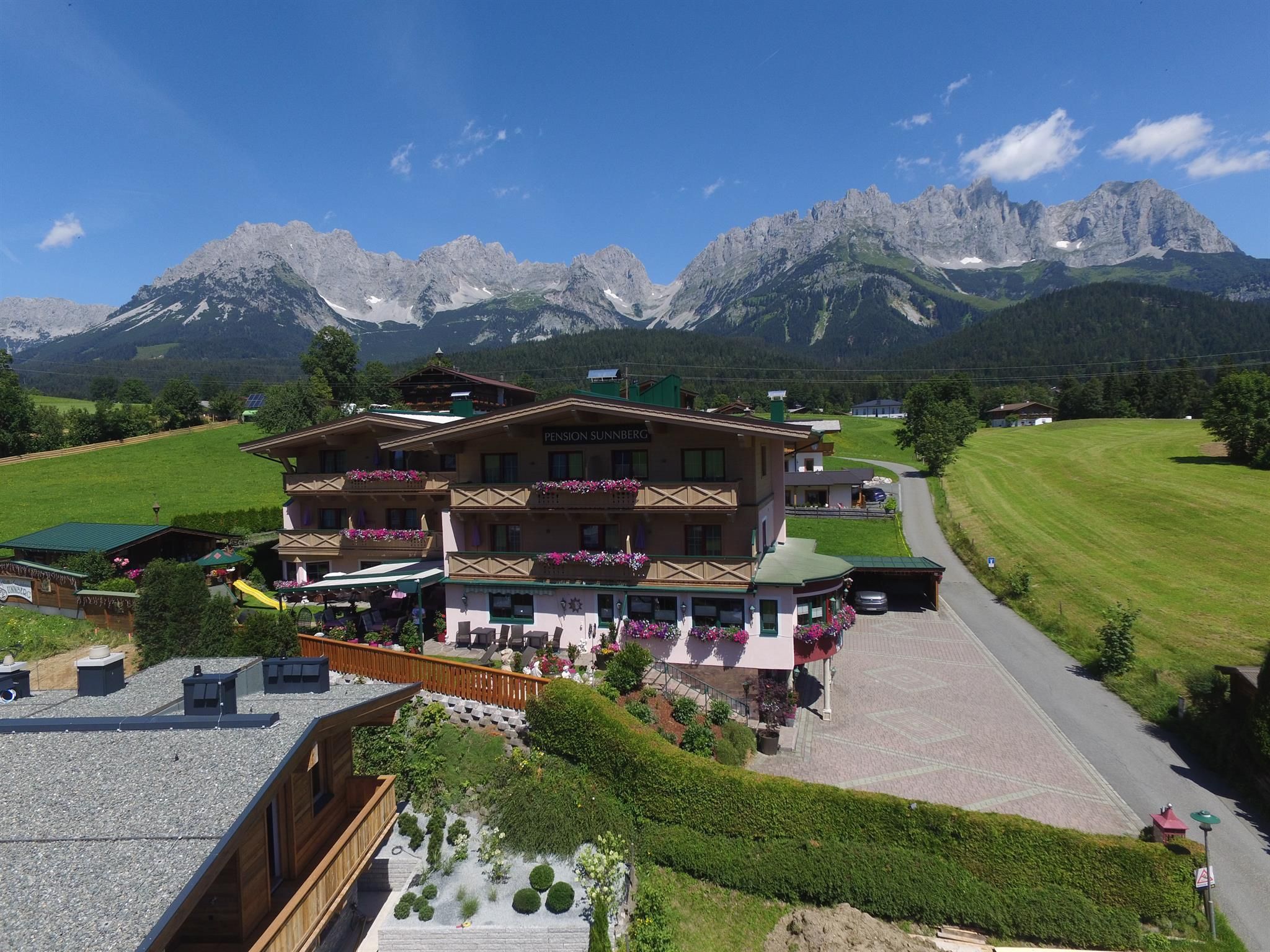 Ein charmantes Chalet in den Alpen mit blühenden Balkonen. Im Hintergrund erheben sich majestätische Berge unter einem klaren blauen Himmel.