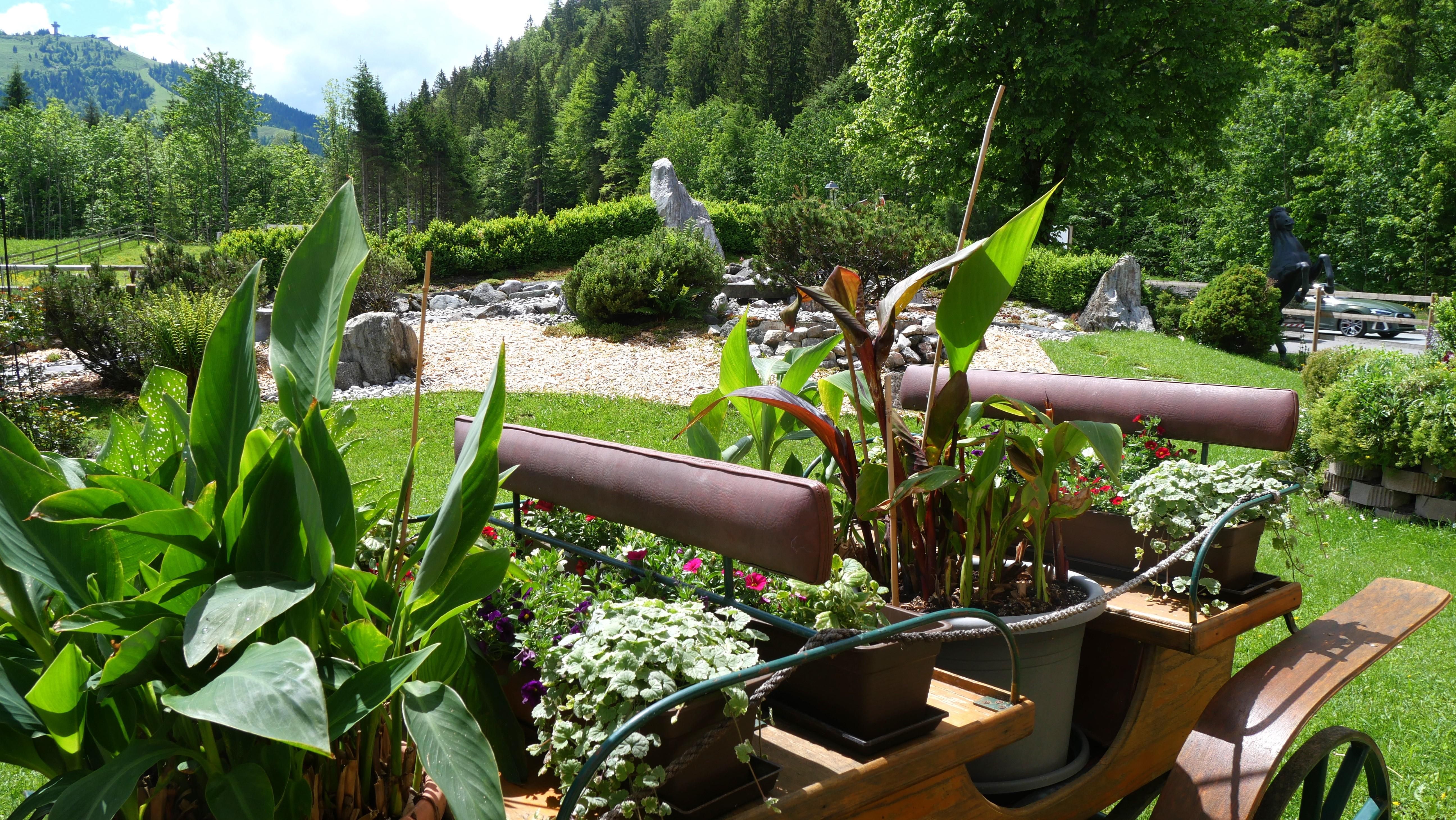 Eine stilvolle Gartenlandschaft mit einem Holzgefährt, das mit Blumen und Pflanzen dekoriert ist. Im Hintergrund sind grüne Bäume und ein kleiner Skulpturengarten zu sehen.