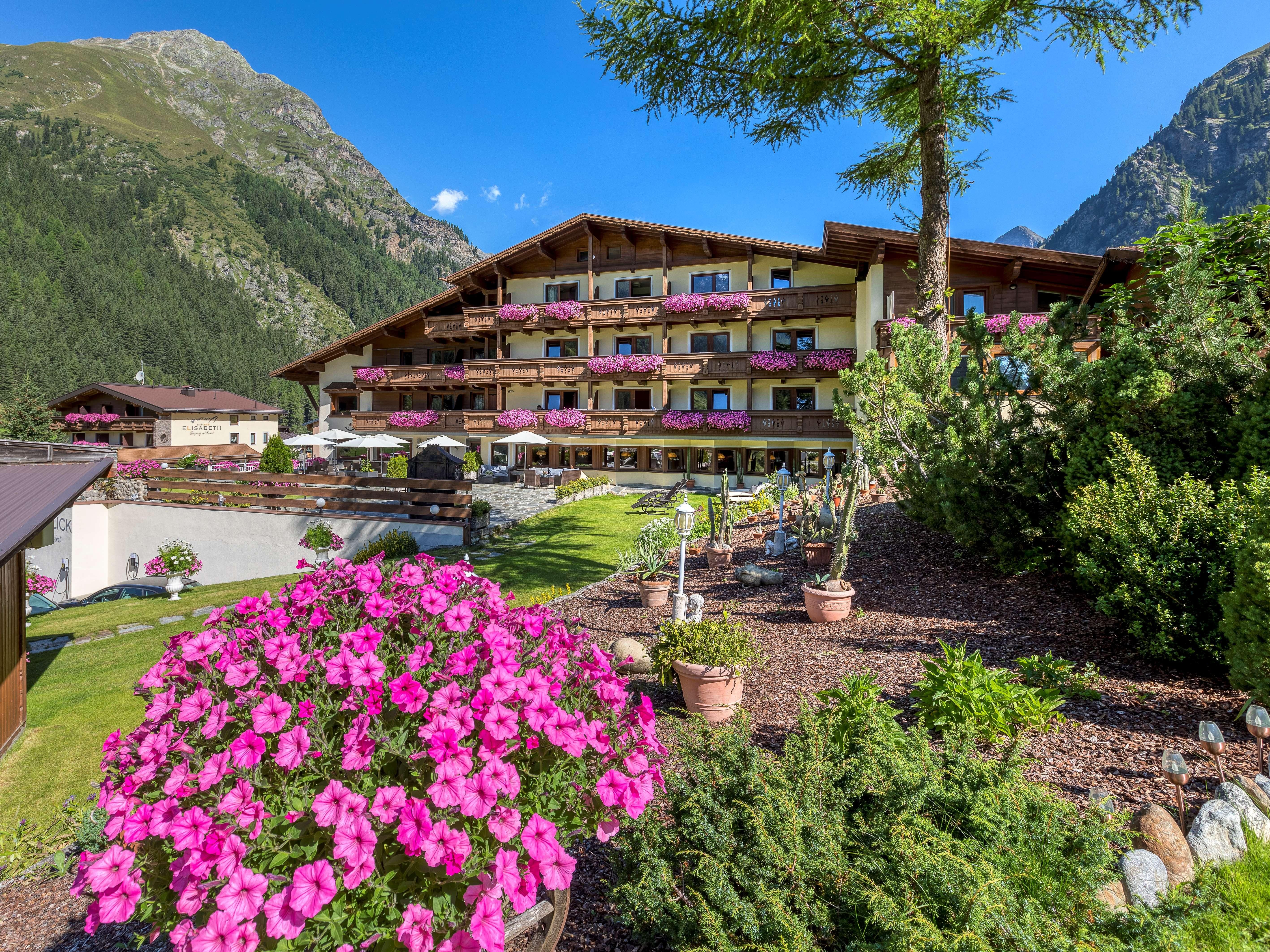 Ein Hotelgebäude umgeben von grünen Bergen und bunten Blumen. Auf den Balkonen blühen pinke Pflanzen und die Umgebung ist einladend.