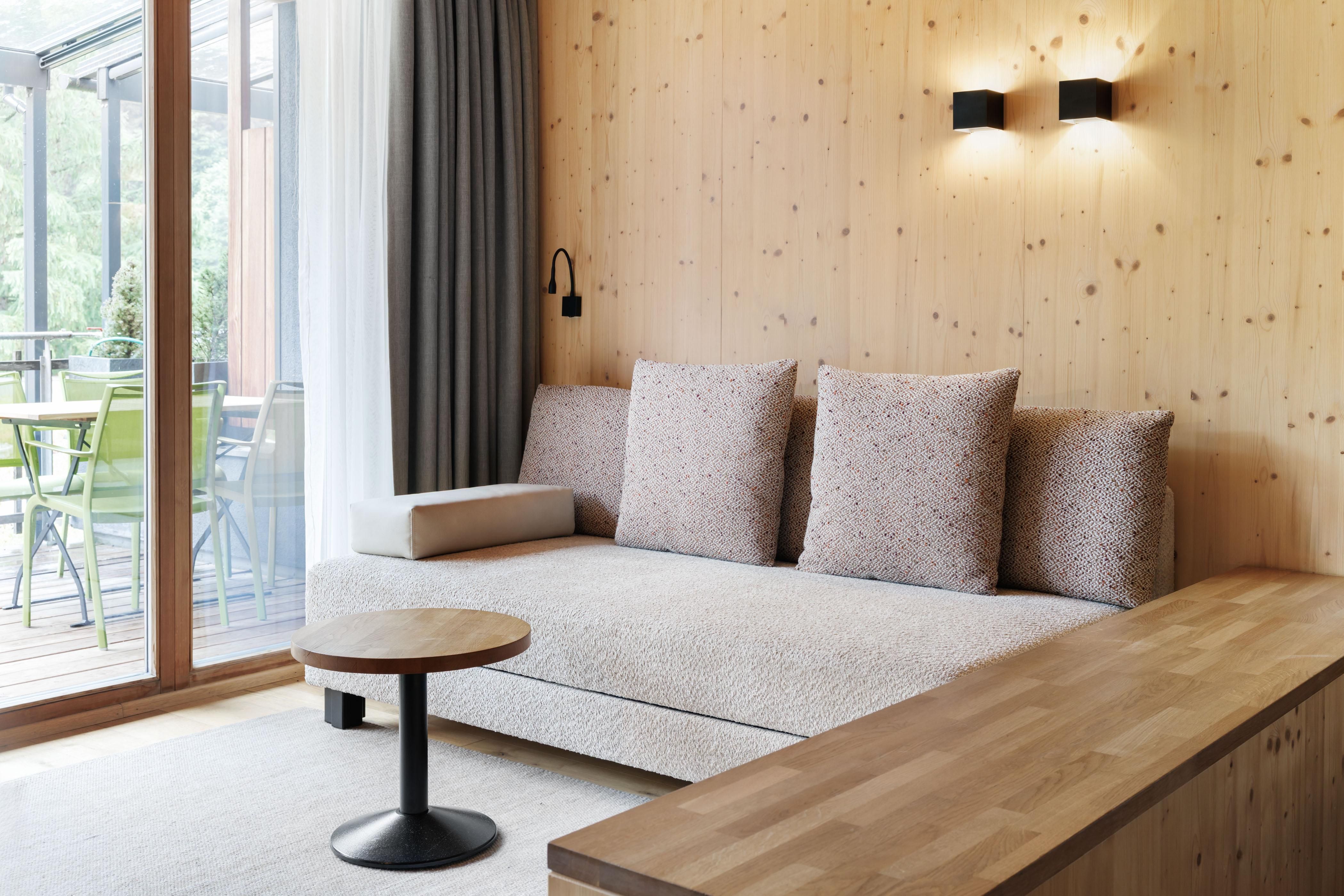 Ein modernes Wohnzimmer mit einem beigen Sofa und großen Kissen. Der Raum hat Holzverkleidung und große Fenster, die auf einen Balkon mit Stühlen blicken.