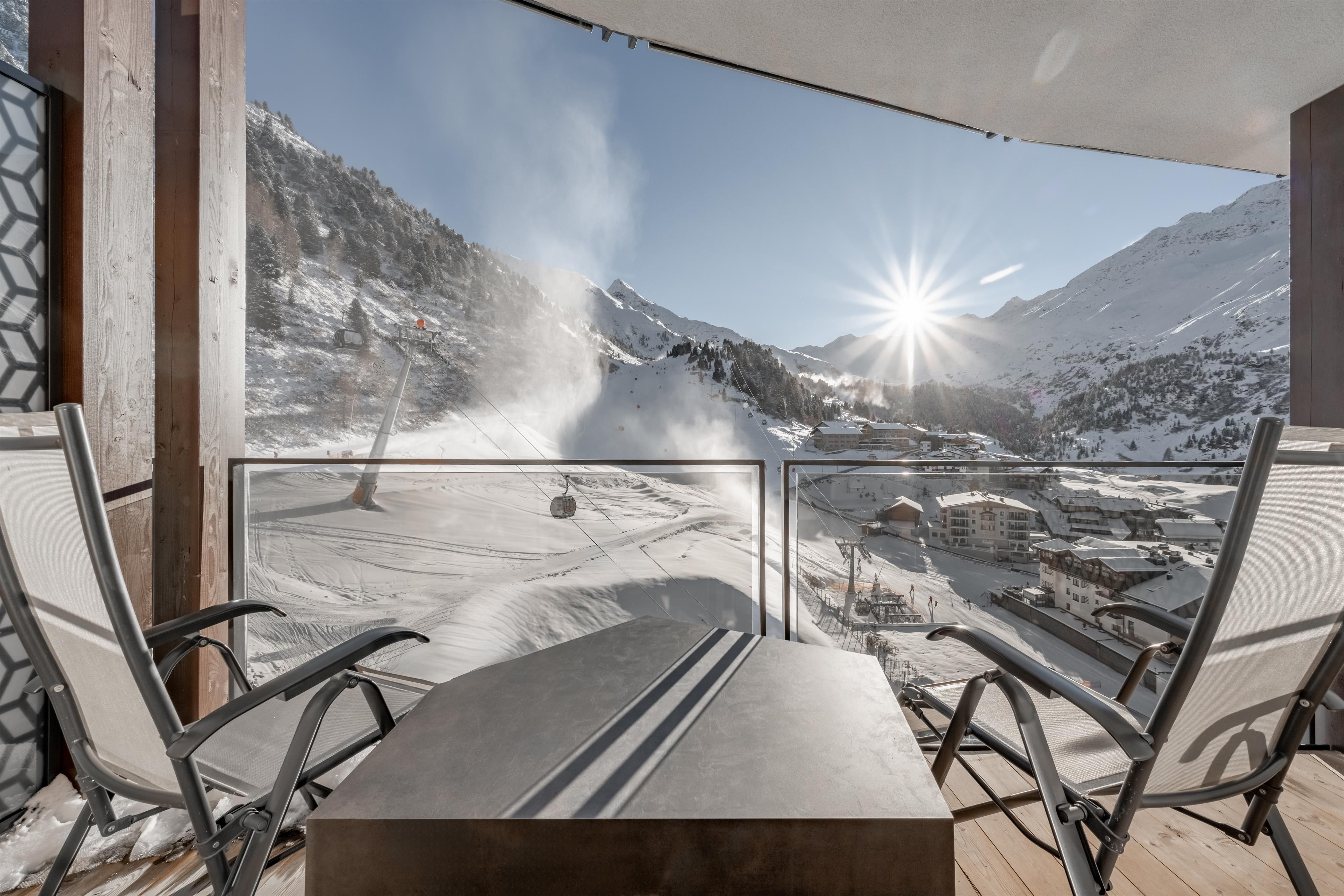 Eine Terrasse mit Stühlen bietet einen Blick auf eine verschneite Berglandschaft. Die Sonne scheint über den Bergen und es gibt eine Skiliftstation in der Nähe.