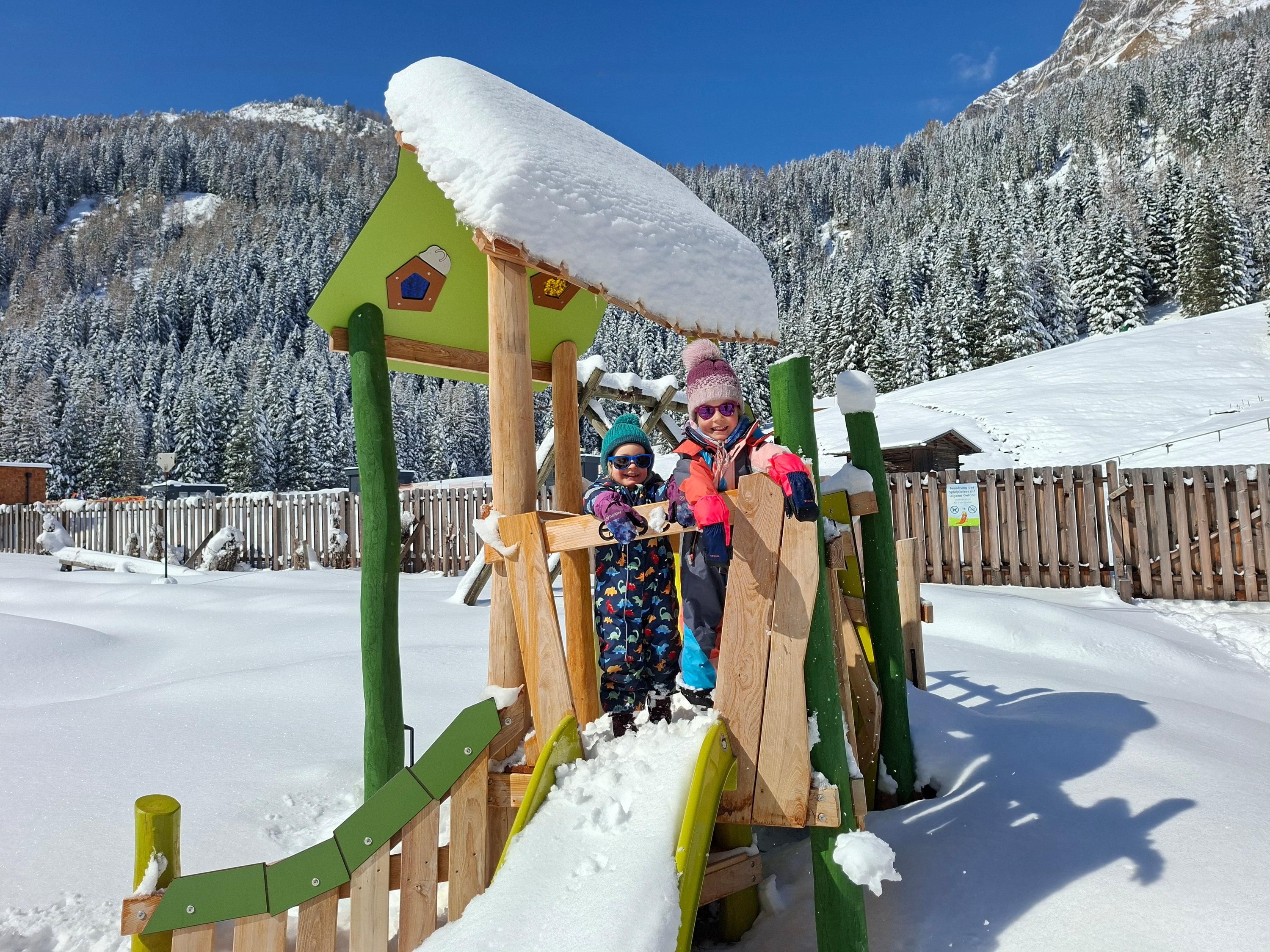 Zwei Kinder spielen auf einem Spielplatz im Schnee. Im Hintergrund sind schneebedeckte Berge und ein blauer Himmel zu sehen.