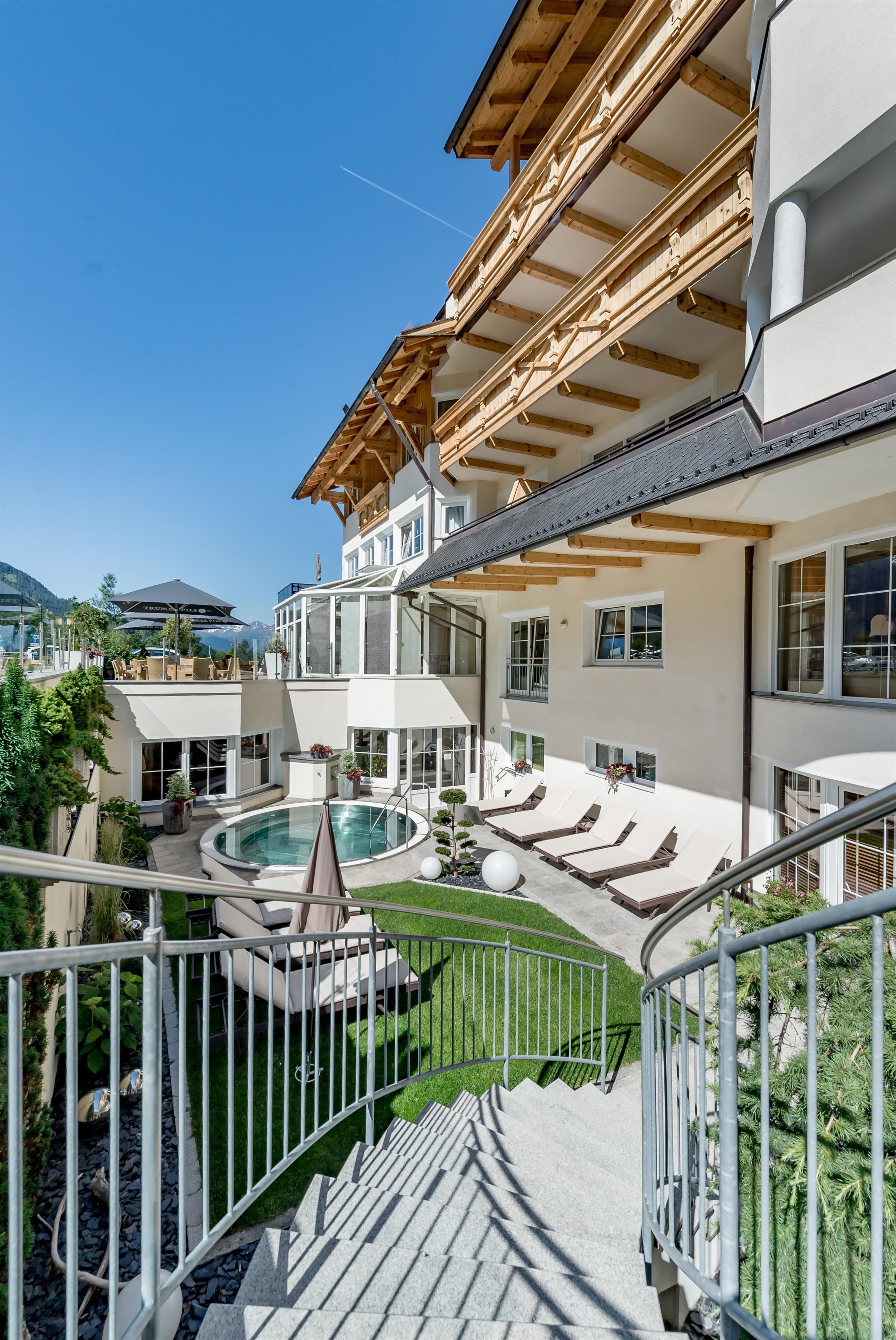 Ein modernes Hotelgebäude mit Holzdetails und einem einladenden Garten. Im Vordergrund ist ein Poolbereich mit Liegestühlen.