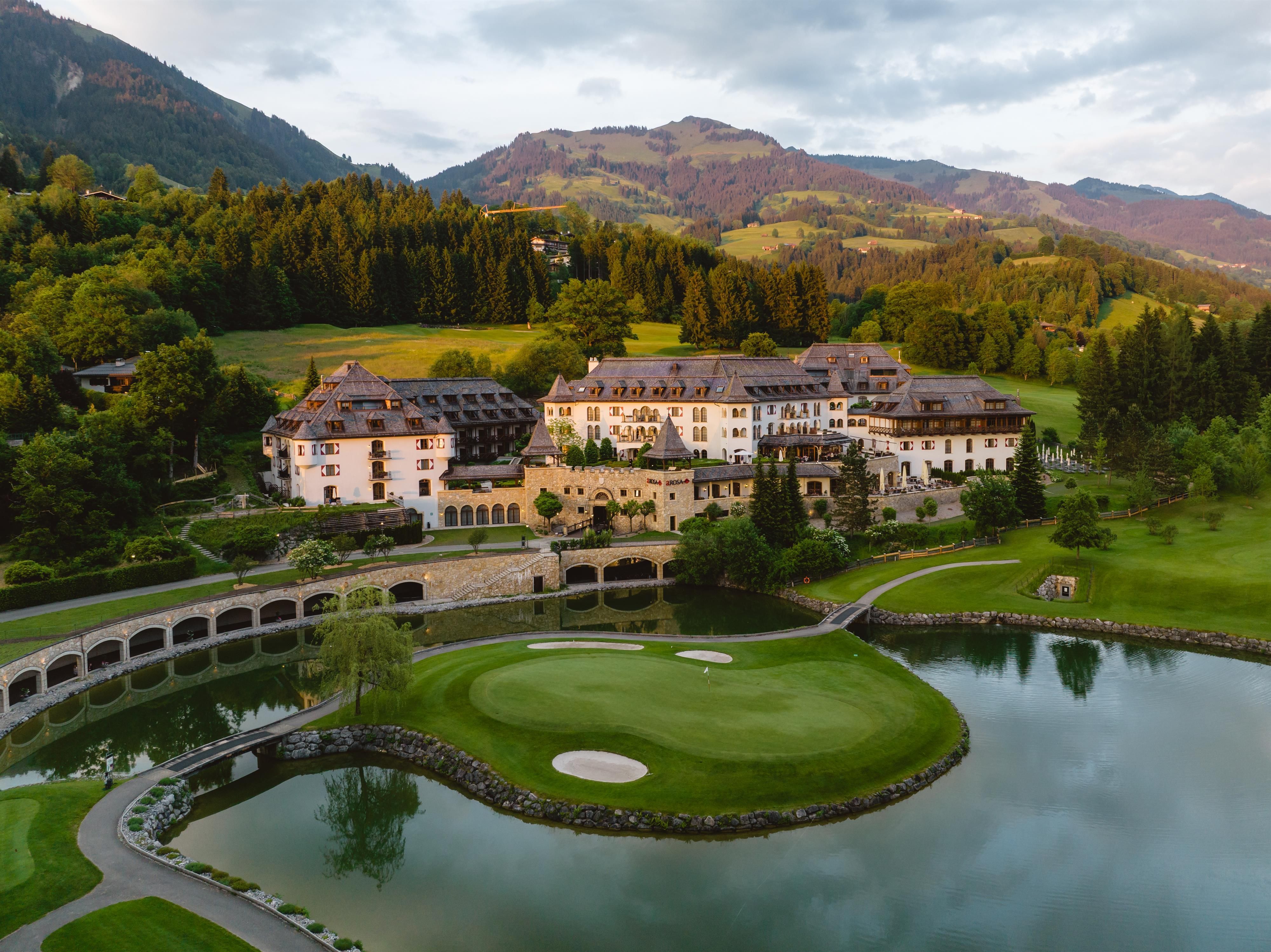 Ein elegantes Hotel inmitten einer grünen Landschaft und bergigen Kulisse. Ein Golfplatz und ein ruhiger See umgeben das Anwesen.