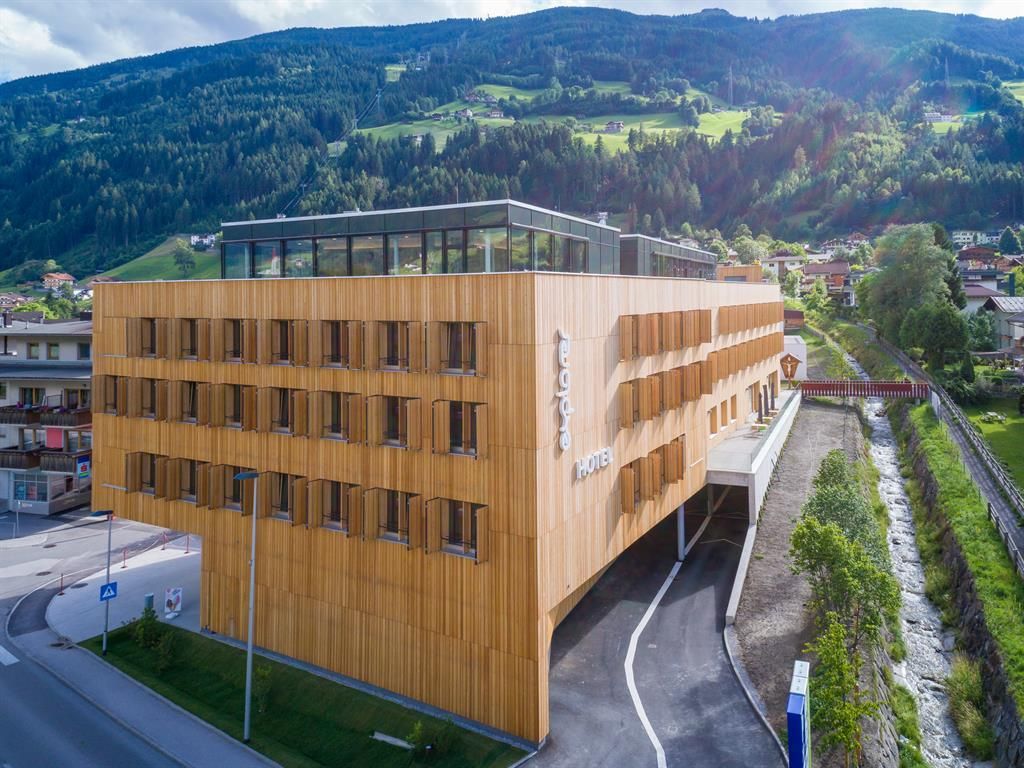 Ein modernes Hotelgebäude aus Holz mit großen Fenstern. Im Hintergrund sind grüne Berge und eine ruhige Straße zu sehen.