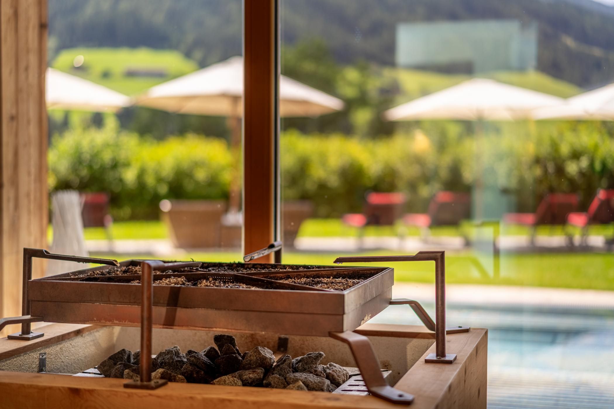 Ein moderne Sauna mit einem Saunaofen aus Holz und Steinen. Im Hintergrund sind eine grüne Landschaft und Liegen mit Sonnenschirmen zu sehen.