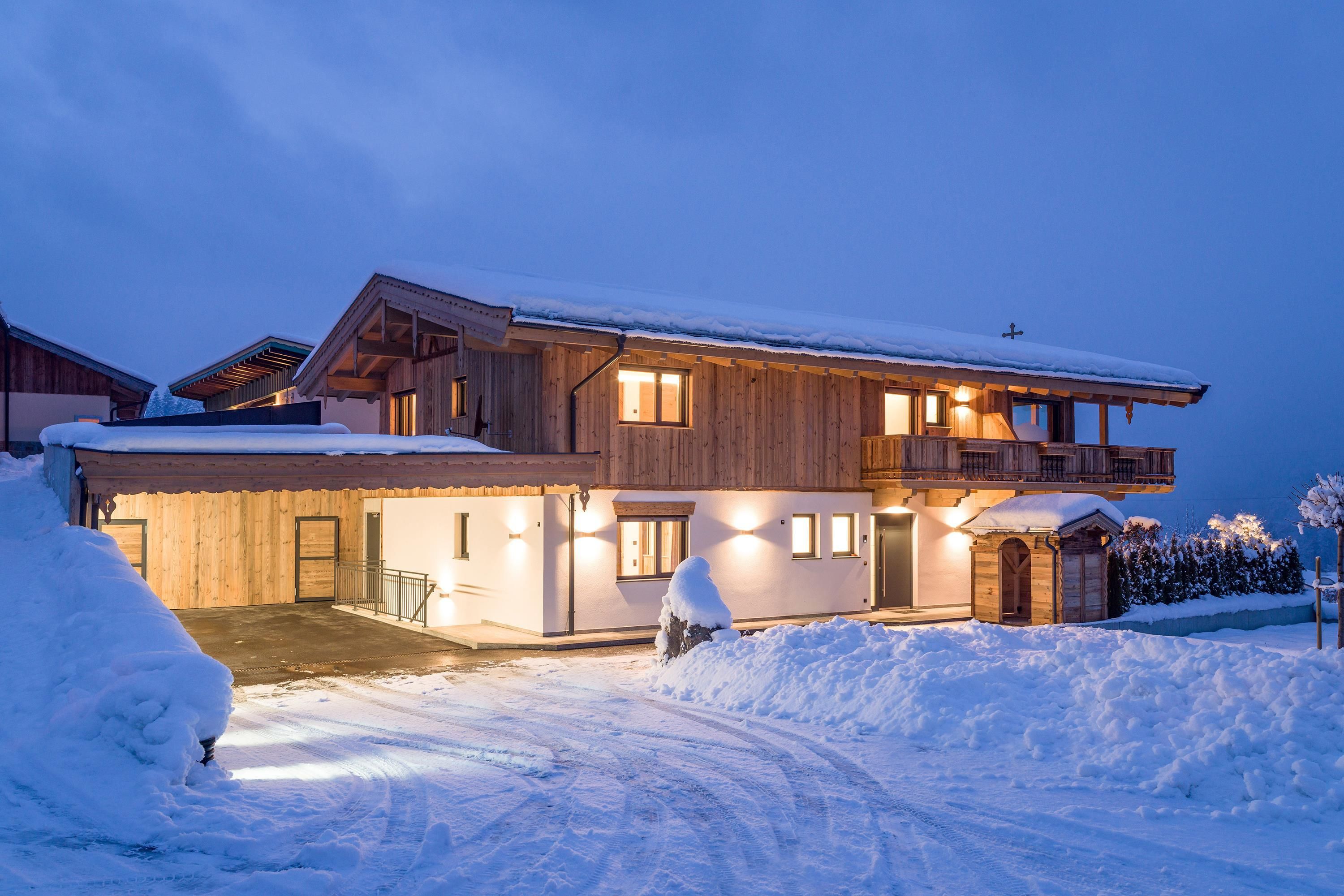 Ein modernes Holzhaus im Schnee, beleuchtet in der Dämmerung. Rundherum liegt frischer, weißer Schnee und der Himmel ist grau-blau.