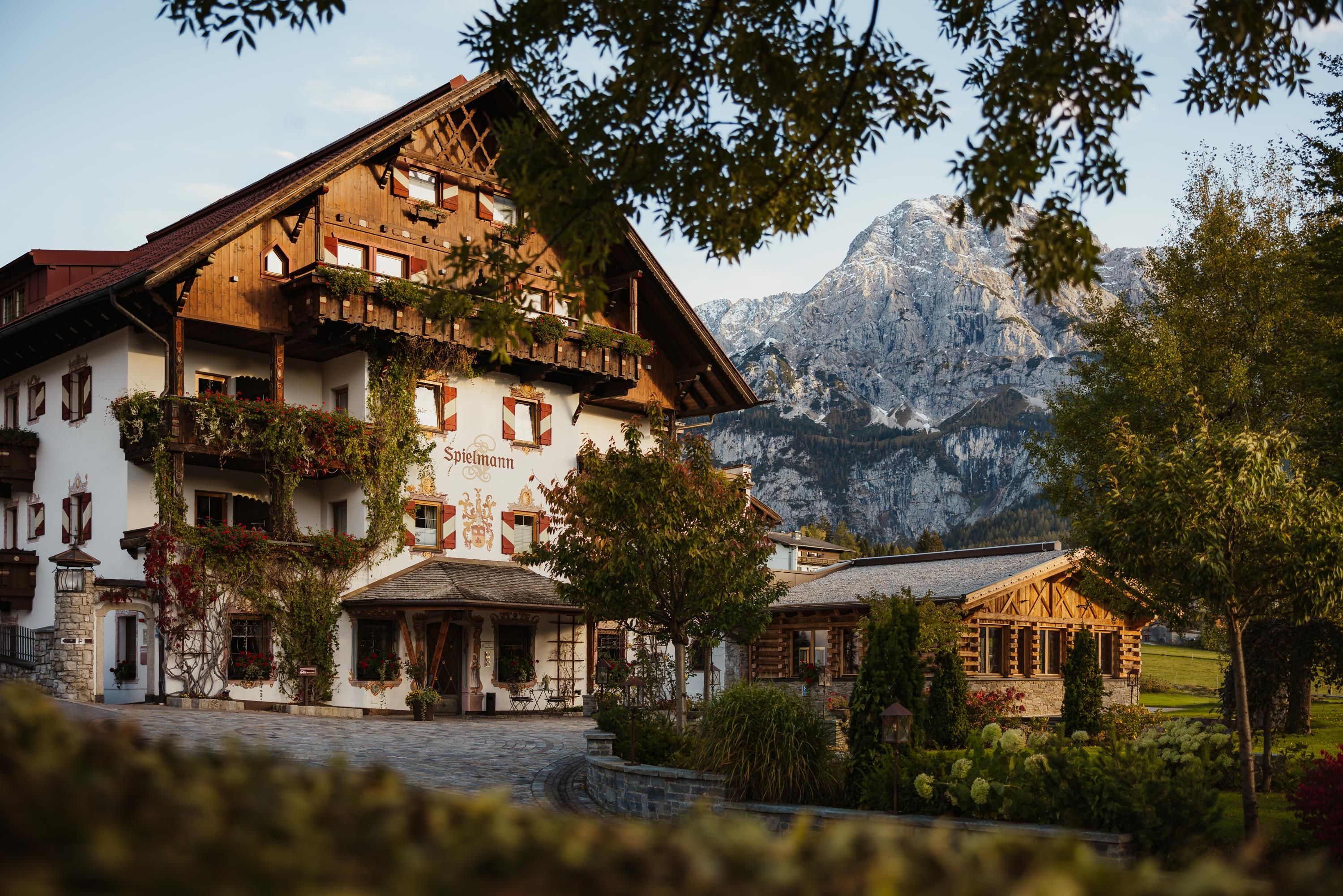 Ein charmantes Bauernhaus in den Bergen, umgeben von Bäumen und grünen Wiesen. Die majestätischen Berge sind im Hintergrund zu sehen.