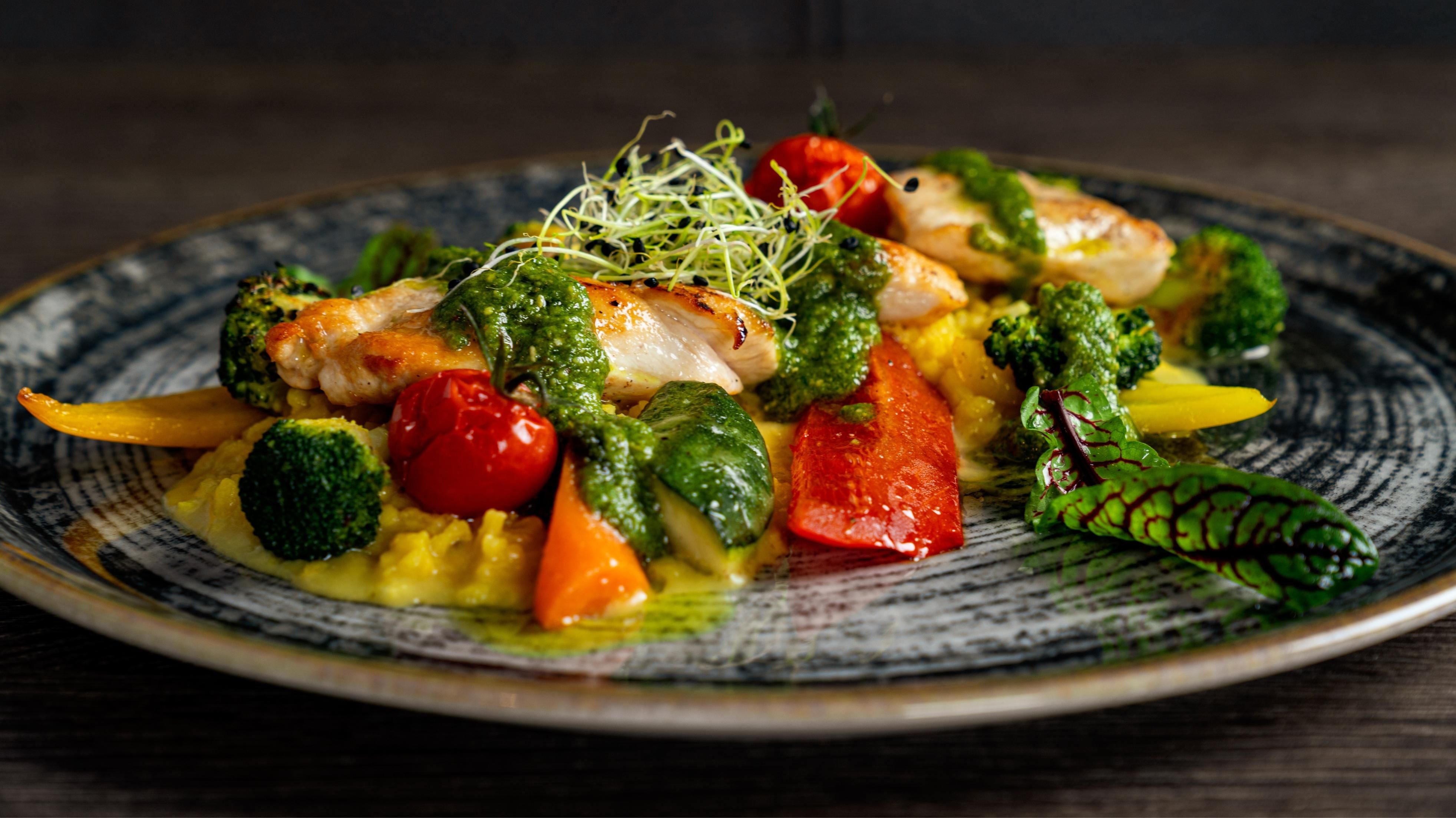 Ein kunstvoll angerichteter Teller mit gebratenem Fisch, buntem Gemüse und einem grünen Pesto. Die frischen Zutaten verleihen dem Gericht eine appetitliche Optik.