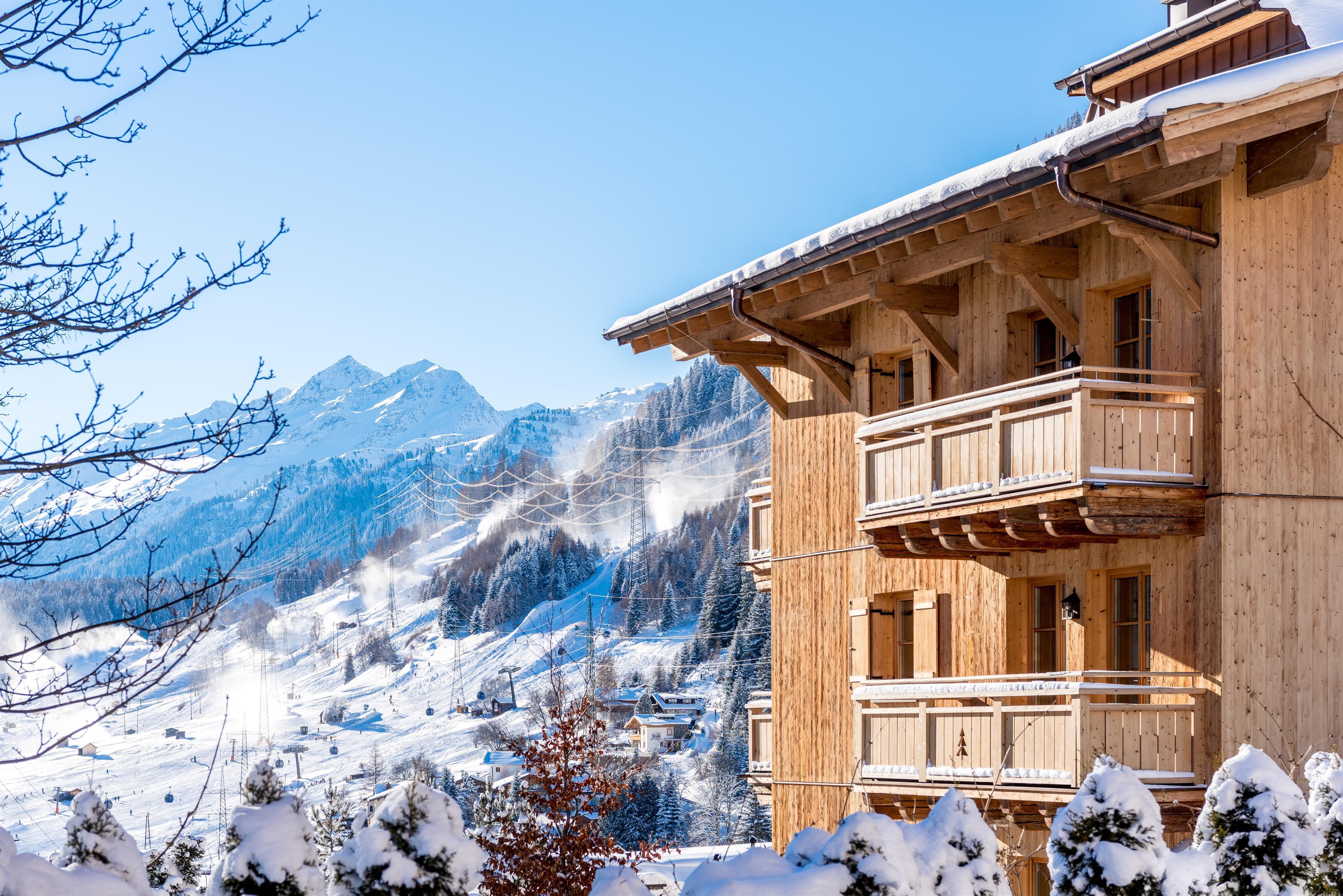 Ein gemütliches Holzgebäude im Schnee mit Balkonen. Im Hintergrund sind schneebedeckte Berge und ein klarer blauer Himmel zu sehen.