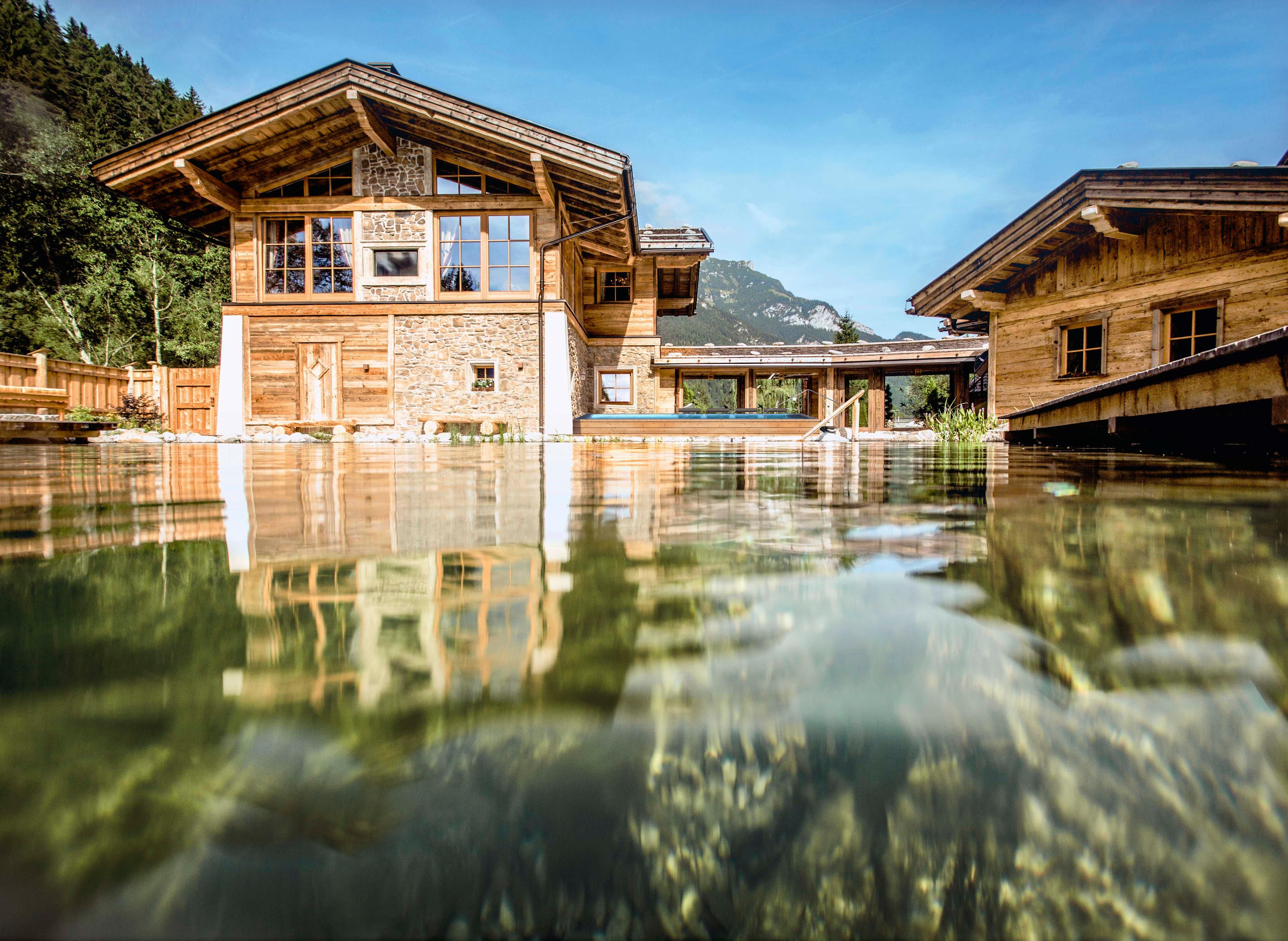 Ein modernes Holzhaus am Wasser mit großen Fenstern und einer schönen Umgebung. Im Hintergrund sind bewaldete Berge zu sehen.