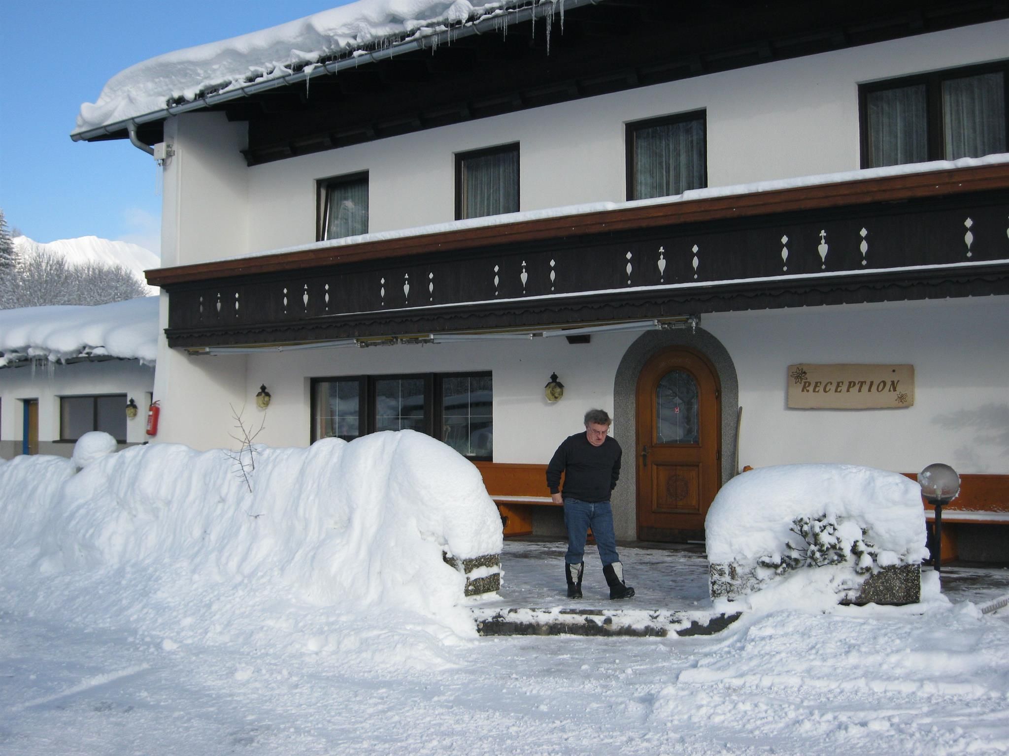 Ein gemütliches Hotel im Schnee mit einem Mann, der vor der Eingangstür steht. Die Umgebung ist winterlich, mit viel Schnee und klarem Himmel.