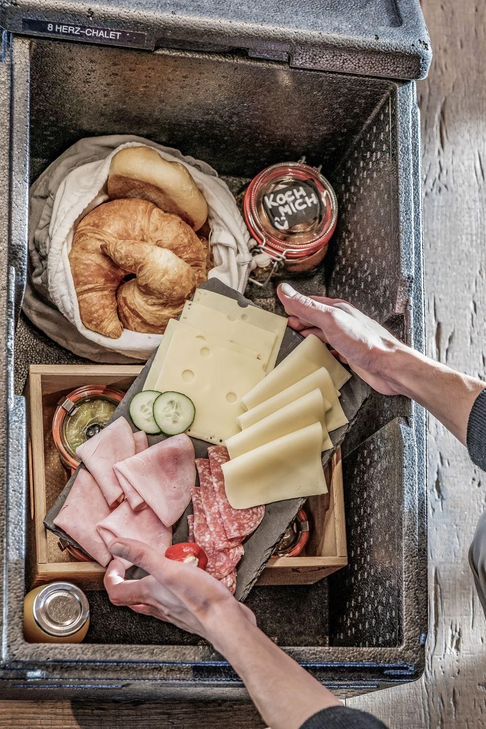 Frühstücksbox voller frischer Lebensmittel, darunter Croissant, Wurst, Käse und Gemüse. Dazu gehören auch Gläser mit Konfitüre und ein Getränk.