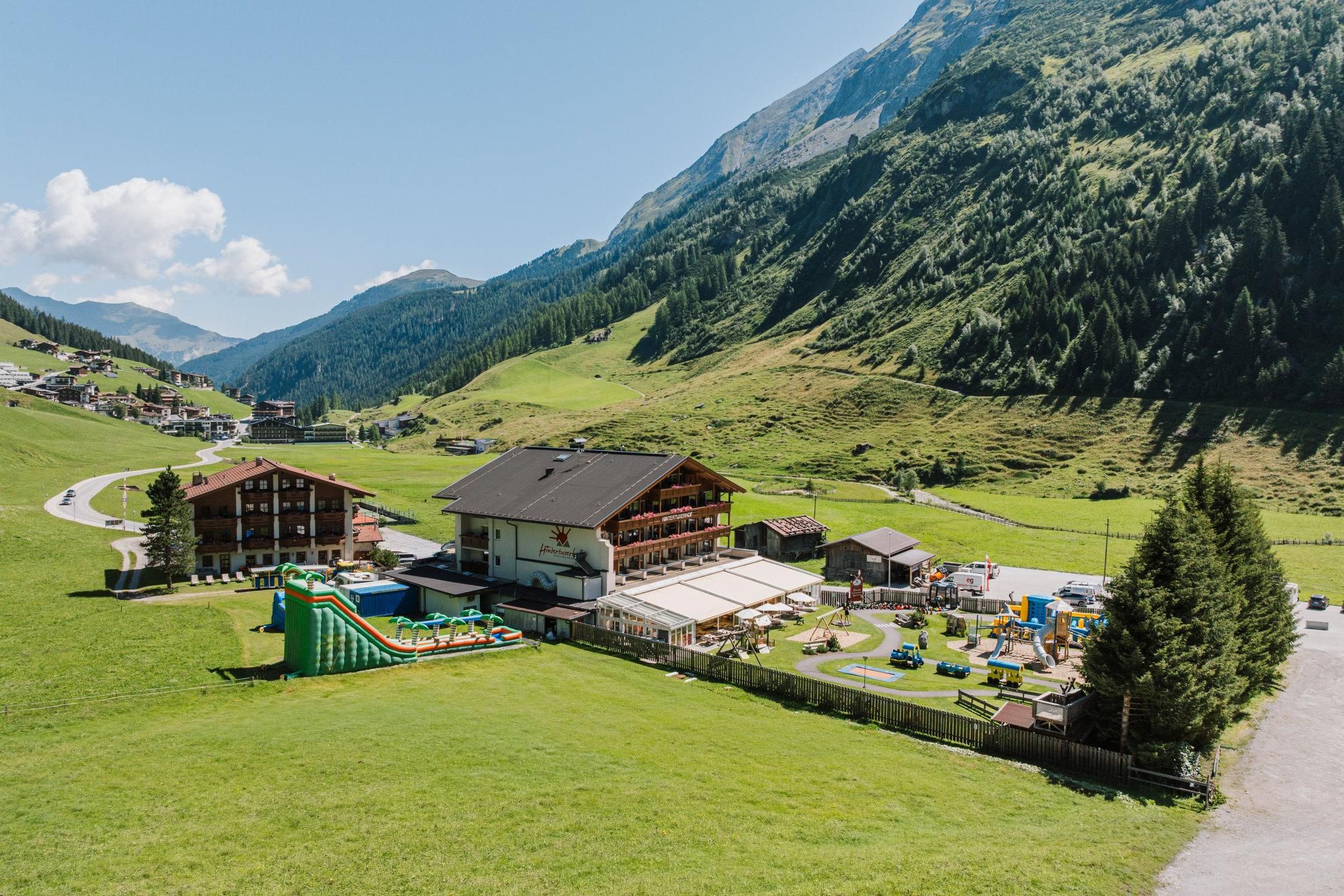 Eine malerische Berglandschaft mit grünen Wiesen und schönen Hütten. Im Vordergrund befindet sich ein Freizeitbereich mit Spielplätzen und Rutschen.