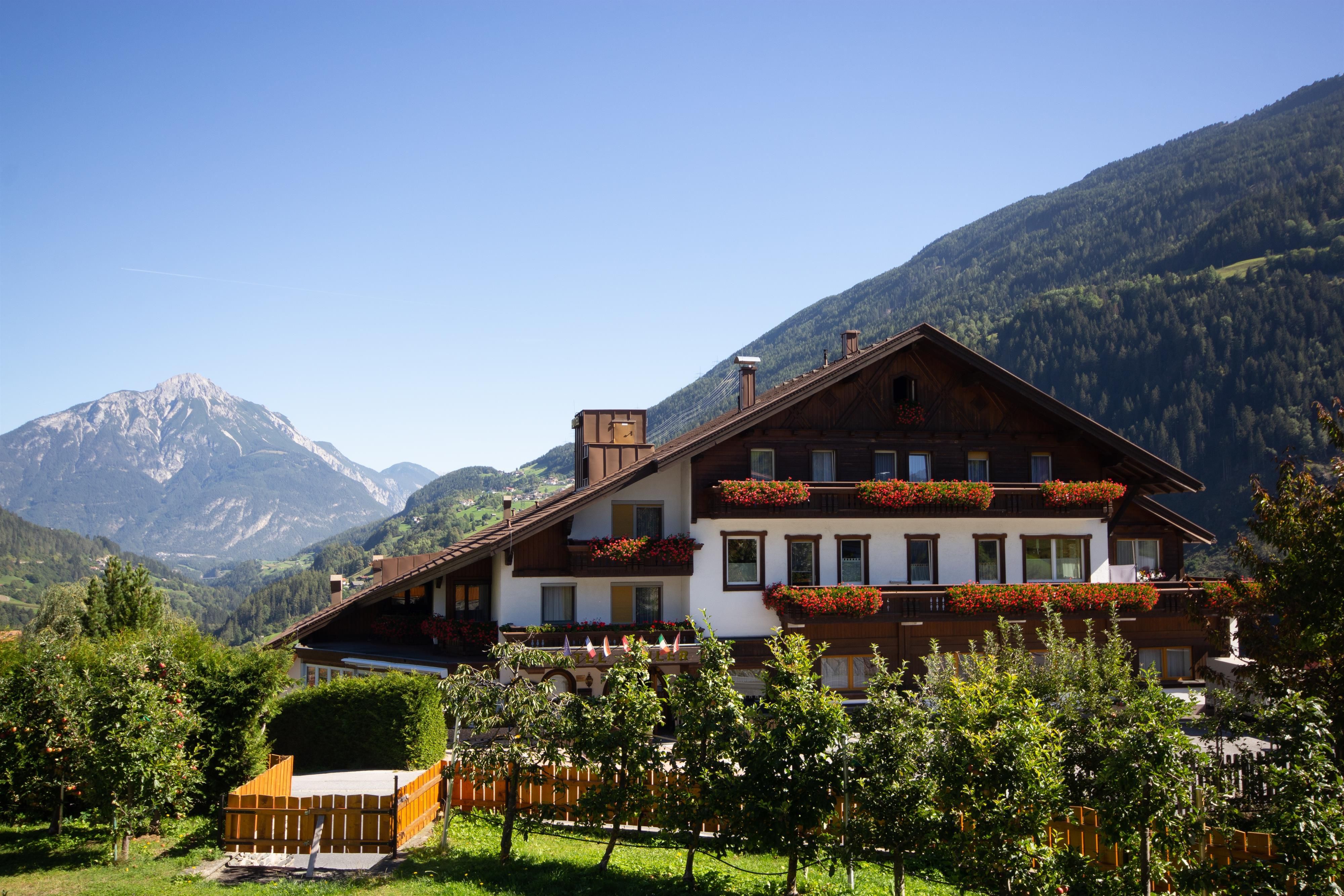 Ein gemütliches Haus in den Bergen mit blühenden Balkonen. Im Hintergrund sind grüne Hügel und schneebedeckte Gipfel zu sehen.