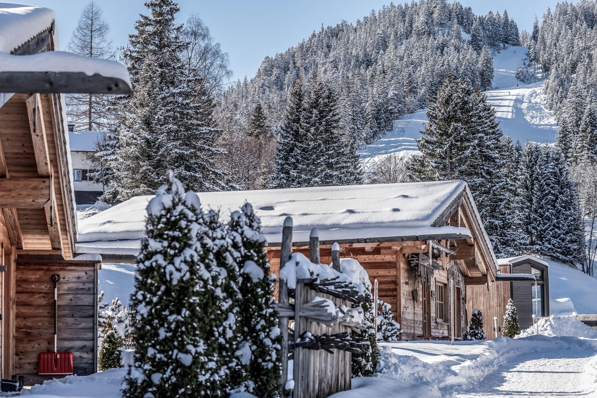 Ein malerisches Winterdorf mit schneebedeckten Chalets und hohen Tannen. Im Hintergrund erkennt man sanfte, verschneite Hügel.