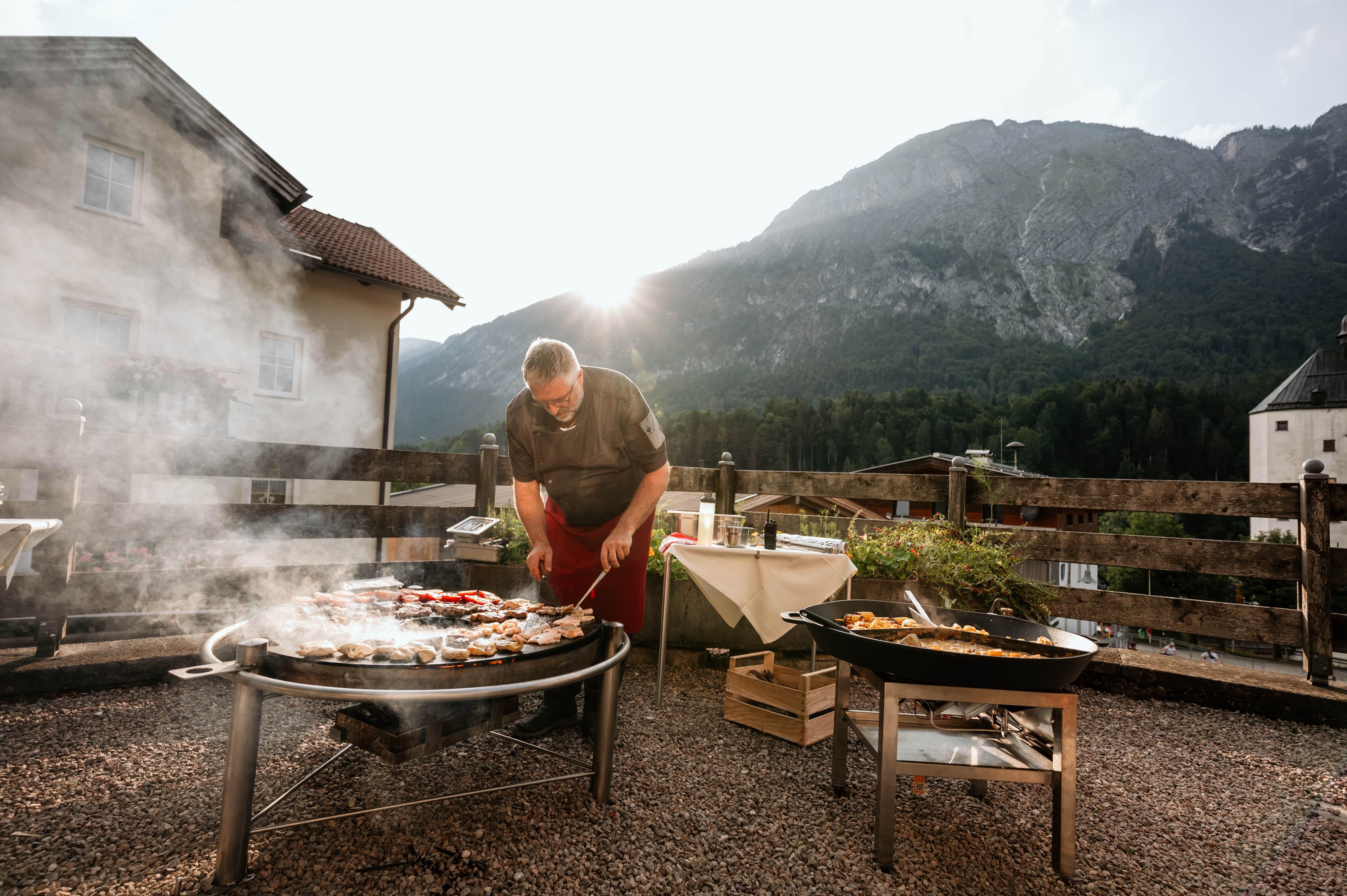 Ein Grillmeister bereitet verschiedene Speisen im Freien zu, umgeben von einer malerischen Berglandschaft. Der Rauch steigt in die Luft, während die Sonne im Hintergrund scheint.