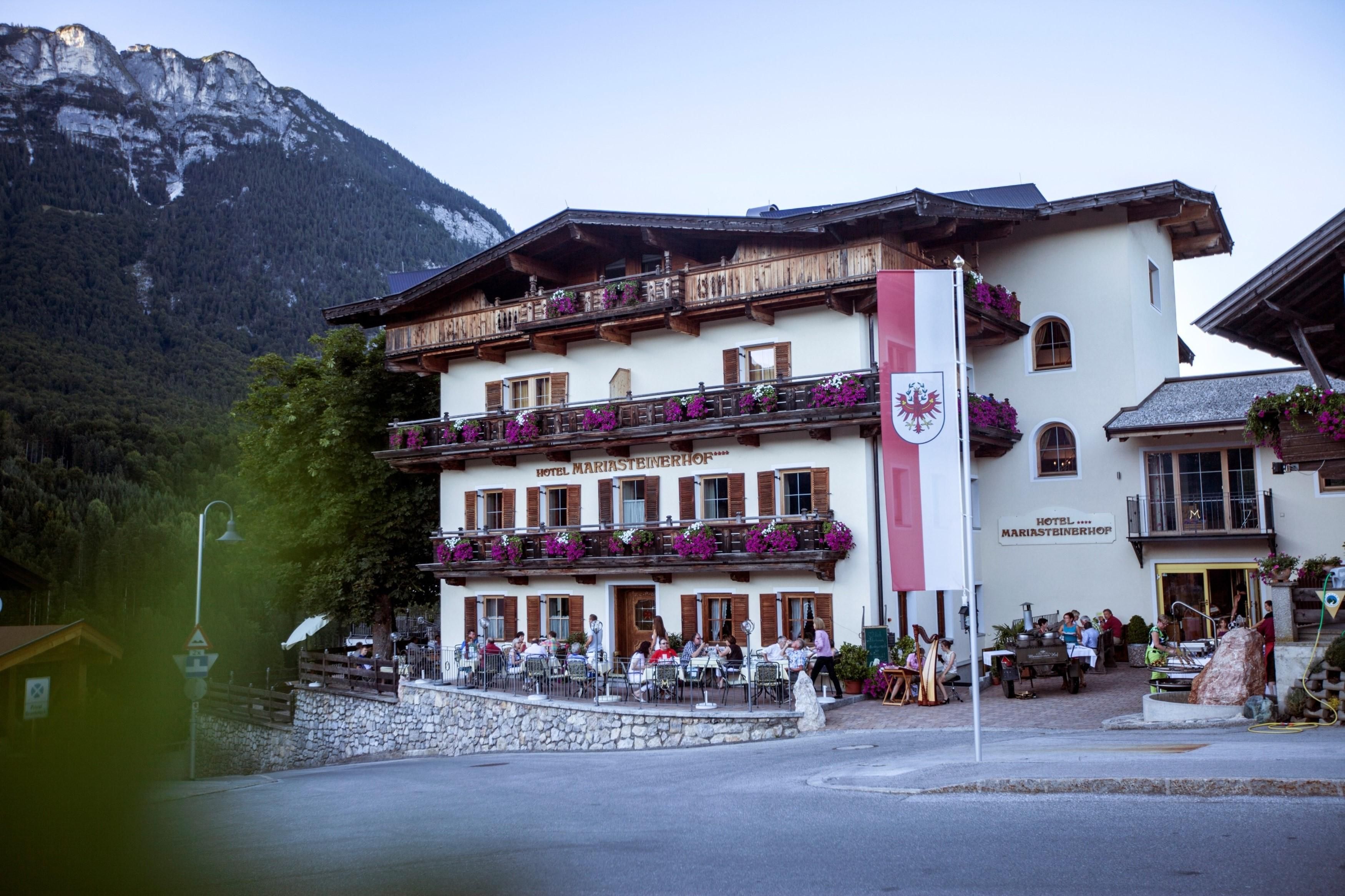Ein gemütliches Hotel in den Alpen mit blühenden Balkonkästen. Im Vordergrund sitzen Gäste auf der Terrasse.