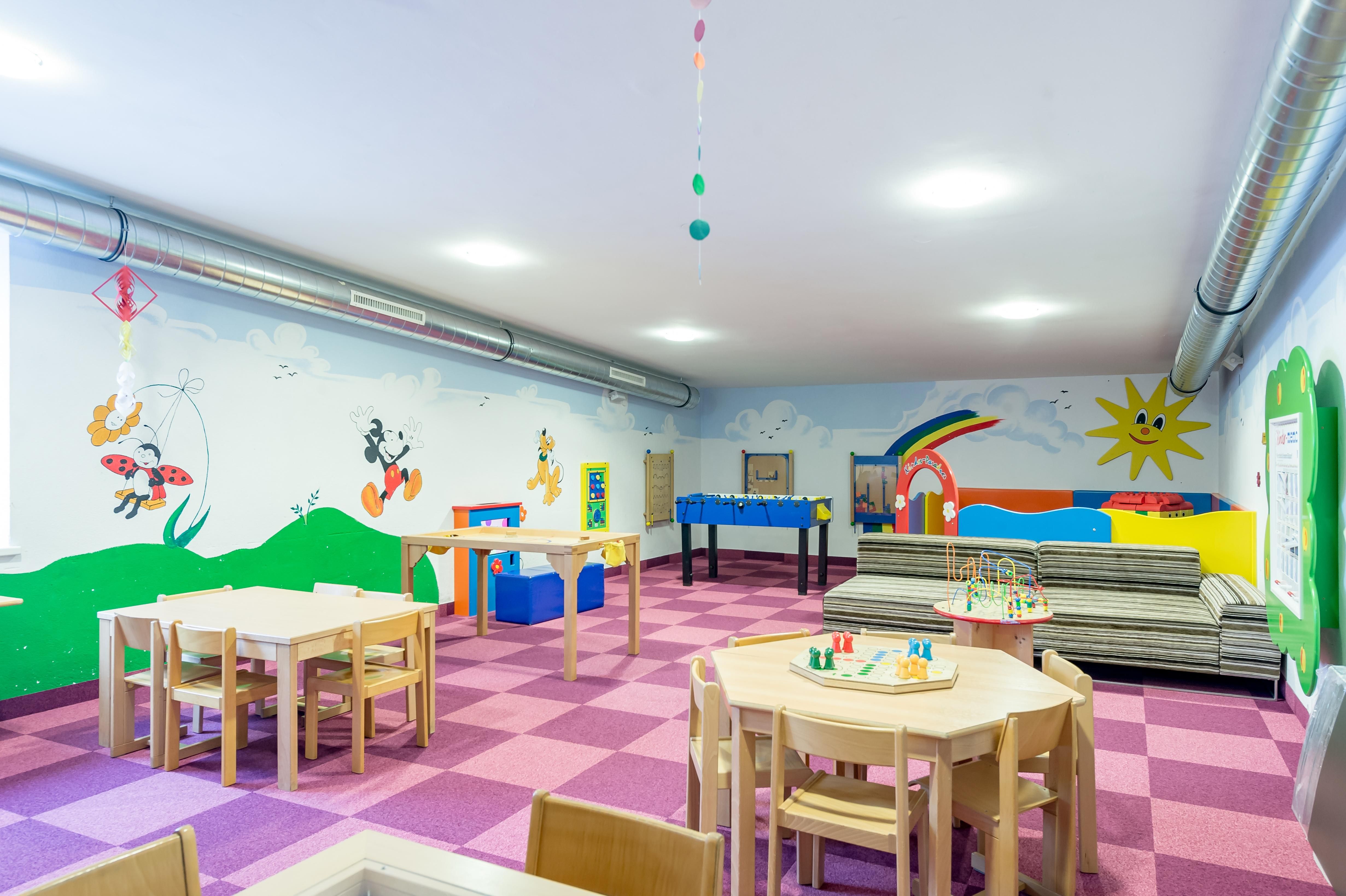 Ein farbenfroher Kindergartenraum mit Tischen und Stühlen. An den Wänden sind bunte Illustrationen und Spielflächen zu sehen.