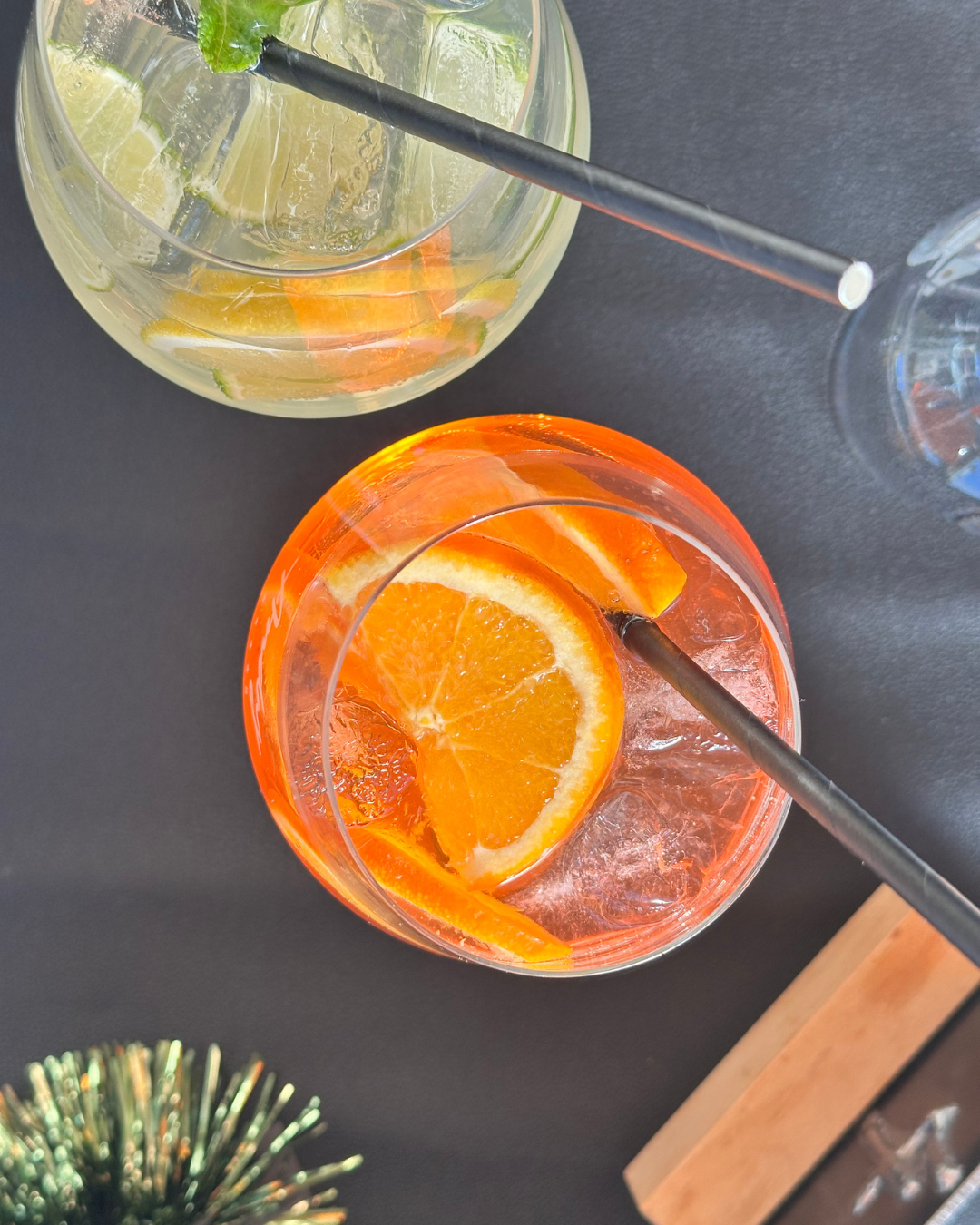 Zwei Cocktails auf einem Tisch, einer mit orangefarbener Flüssigkeit und einer mit einer limonadigen Mischung. Beide Getränke sind mit Eis und frischen Früchten dekoriert.