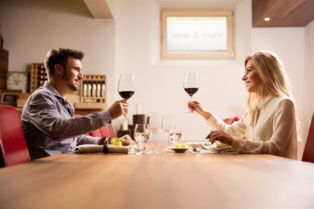 Ein Paar sitzt an einem Tisch in einem Restaurant und prostet sich mit Weingläsern zu. Der Tisch ist mit Speisen und weiteren Gläsern gedeckt.