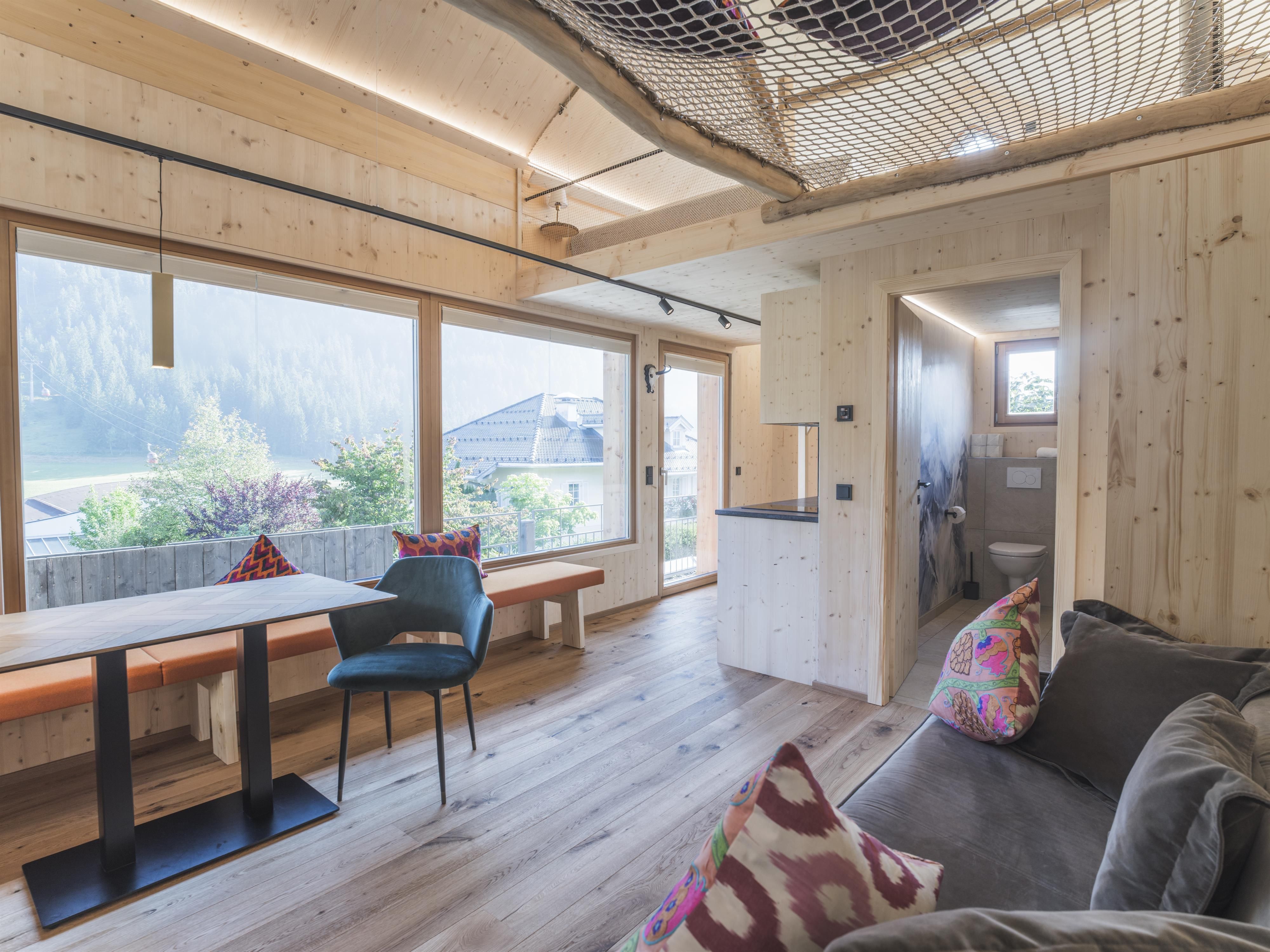 Ein modernes Wohnzimmer mit hellem Holzdesign und großen Fenstern. Es gibt einen Tisch mit Stühlen und einen gemütlichen Bereich mit einem Sofa und bunten Kissen.