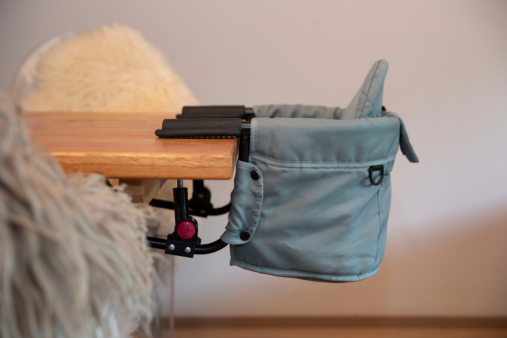 Ein hochstuhlähnlicher Sitz, der an einem Tisch befestigt ist. Der Sitz hat eine weiche Polsterung und eine praktische Tasche an der Seite.