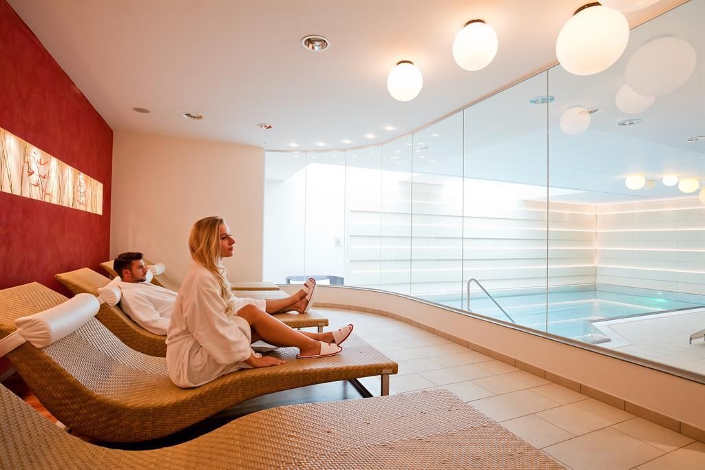 Ein modernes Spa mit komfortablen Liegen und großen Fenstern zum Poolbereich. Ein Mann und eine Frau entspannen in einem ruhigen Ambiente.