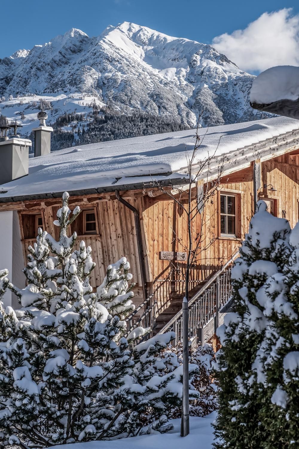 Ein gemütliches  Chalet im Schnee, umgeben von schneebedeckten Bäumen. Im Hintergrund sind majestätische Berge unter einem klaren blauen Himmel sichtbar.
