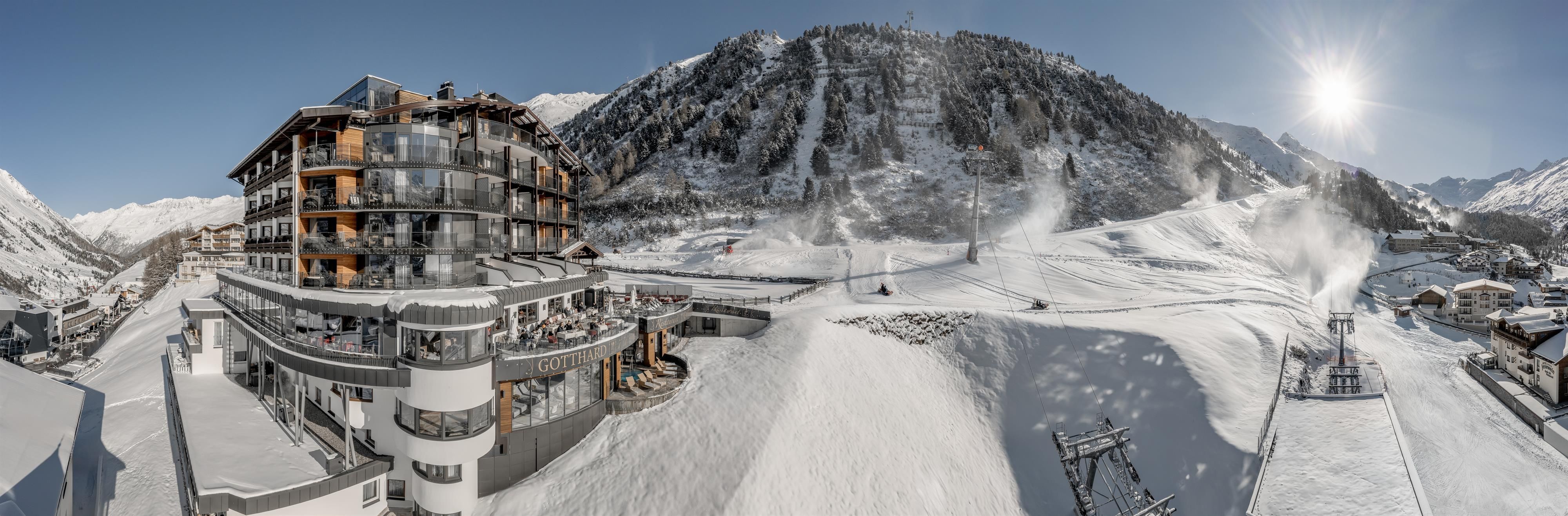 Ein modernes Hotel in einem verschneiten Skiort. Im Hintergrund sind Berge und Skilifte zu sehen.