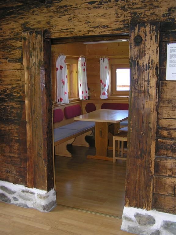 Ein einladendes Zimmer mit Holzvertäfelung und einem großen Tisch. Durch den Türrahmen sind die schlichten, traditionellen Möbel und Fenster mit weißen Vorhängen sichtbar.