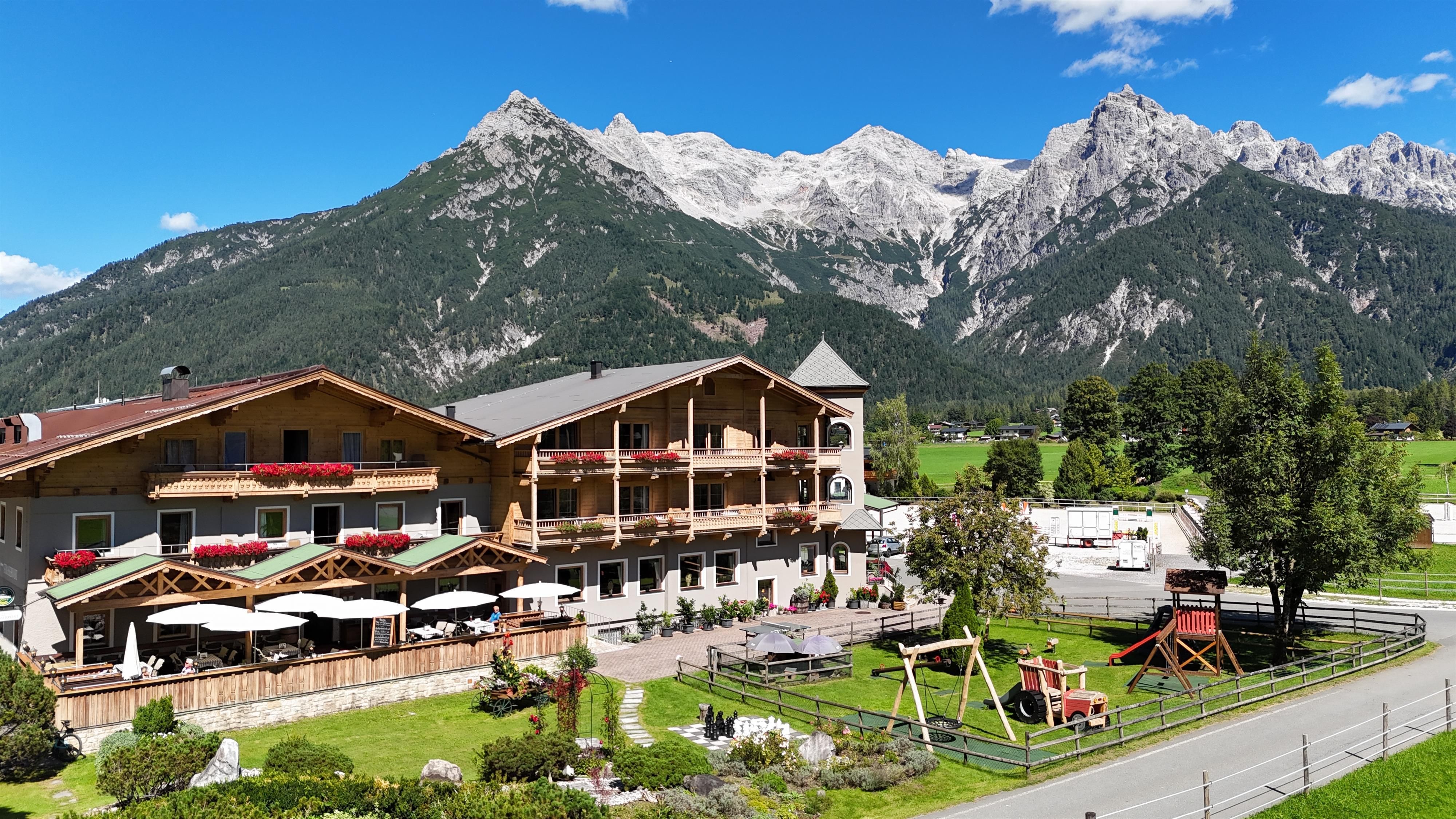 Ein malerisches Hotel mit Terrasse und Spielplatz, umgeben von majestätischen Bergen. Der Himmel ist klar und blau, ideal für einen entspannenden Aufenthalt in der Natur.