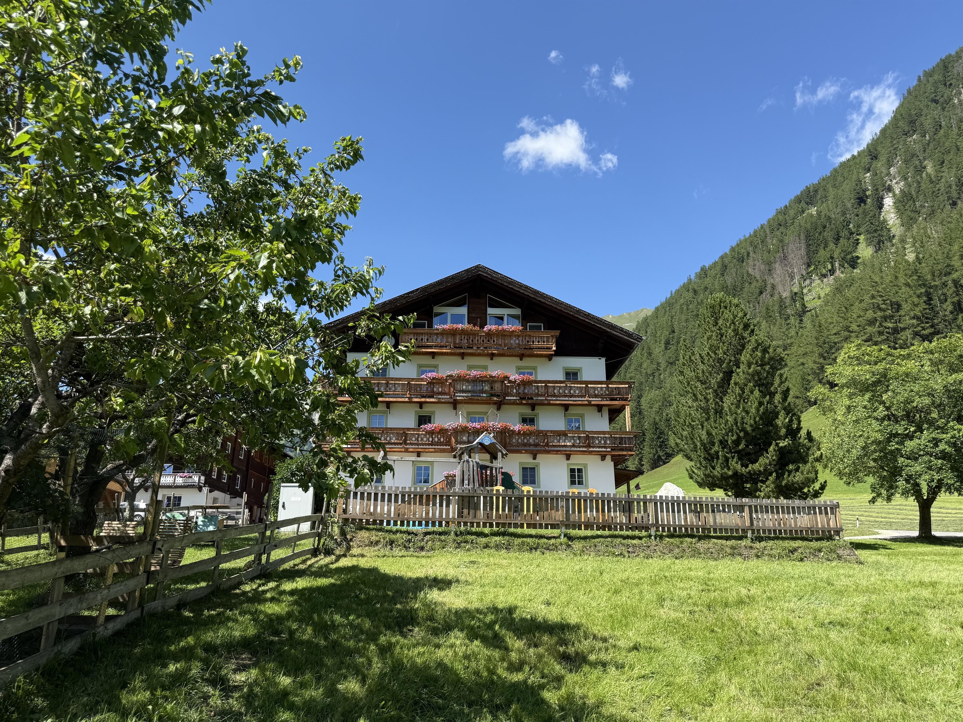 Das Bauernhaus mit Holzbalkone und davor liegt eine grüne Wiese mit Obstbäume