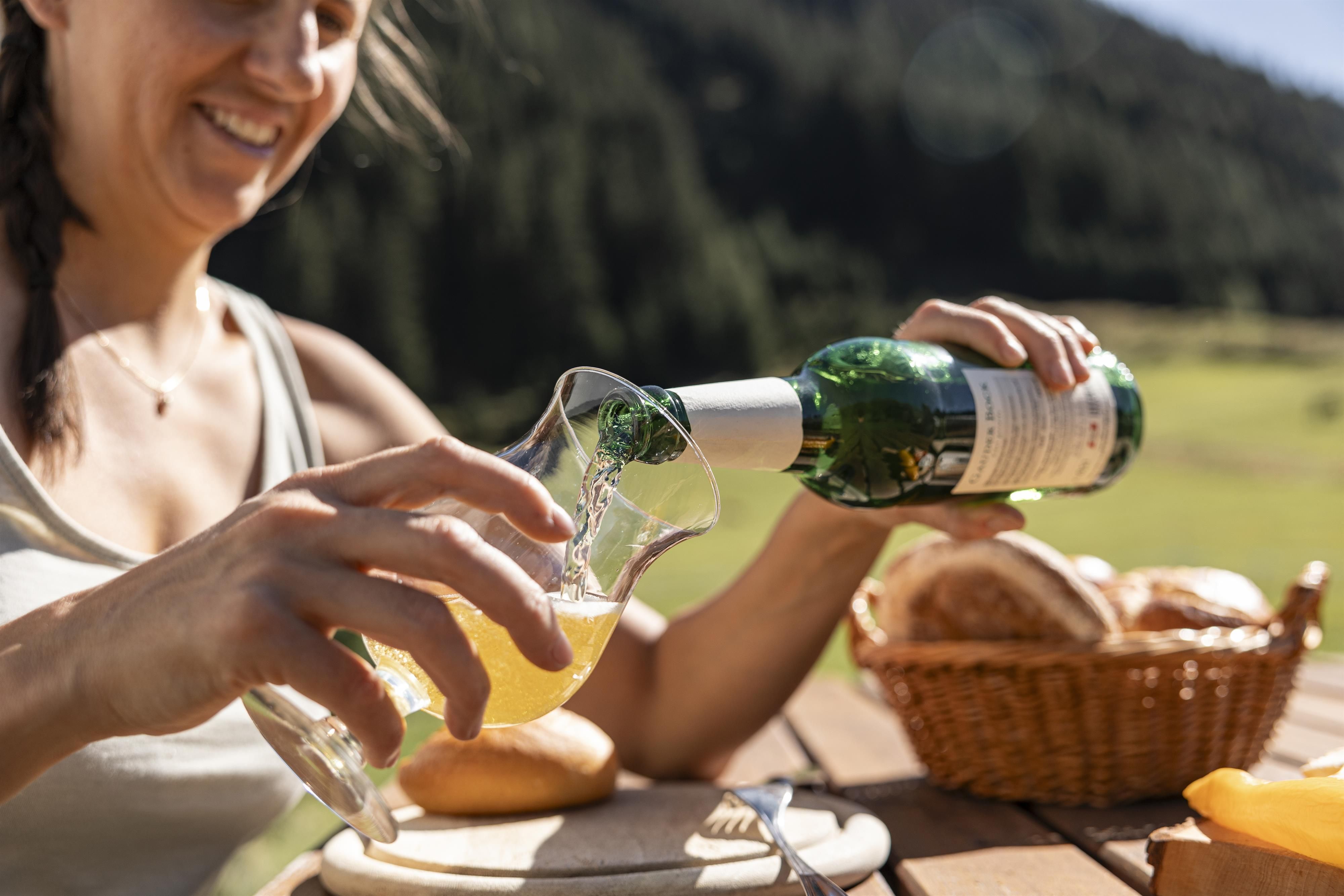 Eine Frau schenkt ein Getränk aus einer Flasche in ein Glas. Im Hintergrund sind eine grüne Landschaft und ein Korb mit Brötchen zu sehen.