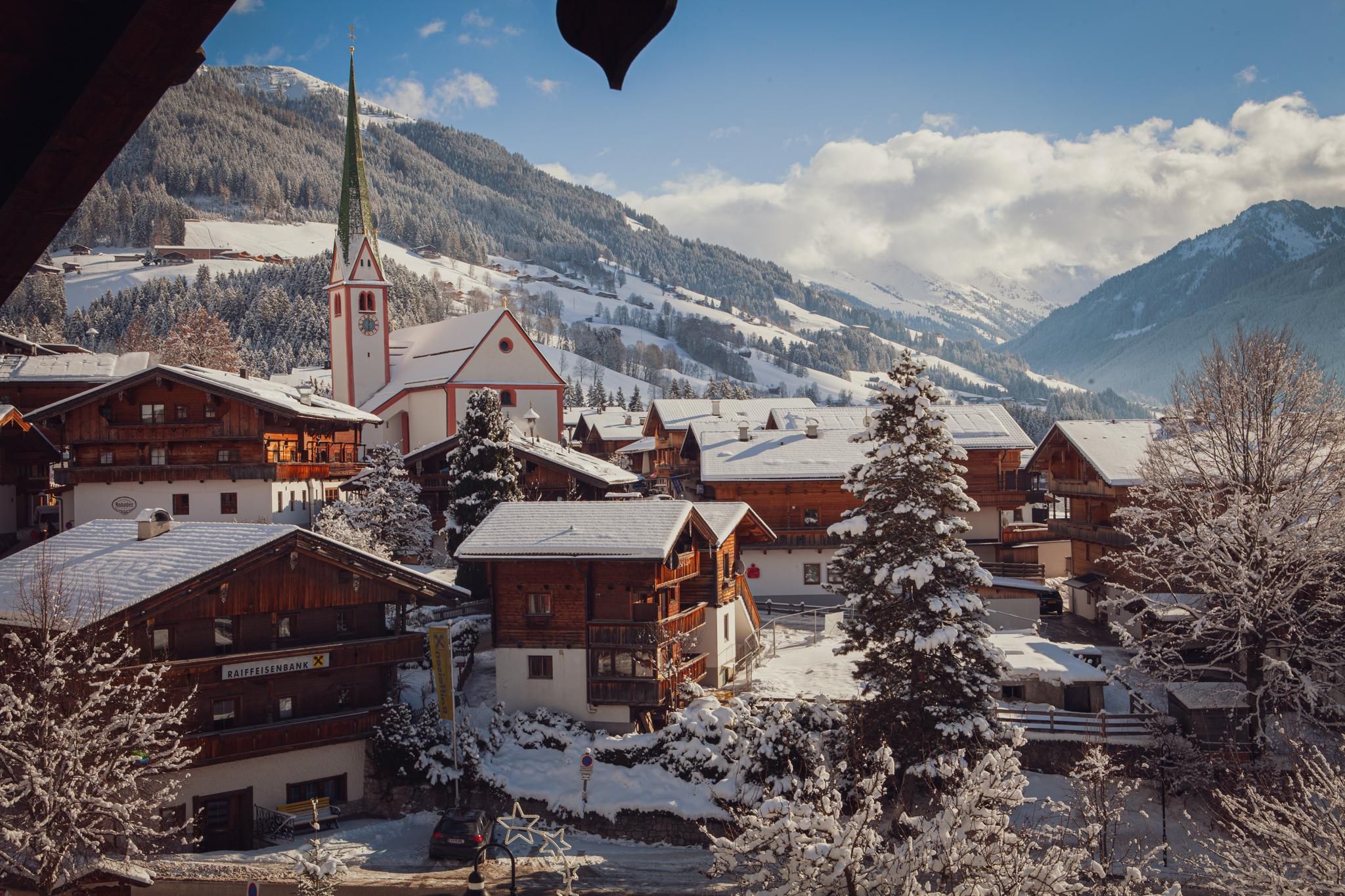 Ein malerisches Dorf im Winter mit schneebedeckten Häusern und einer Kirche. Die umliegenden Berge sind im Hintergrund sichtbar, unter einem klaren Himmel.